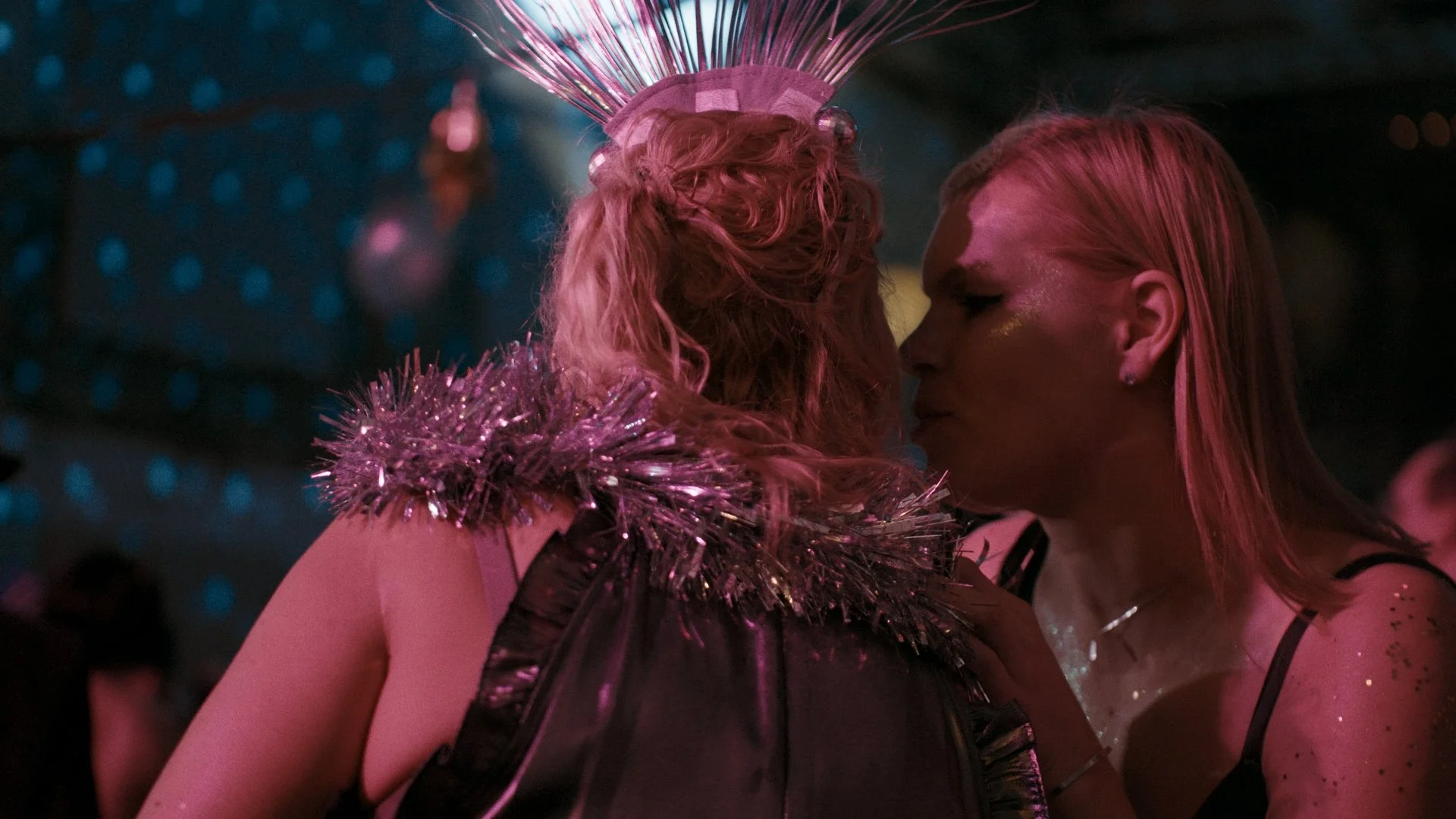 Two women whispering at an event with colorful lighting.