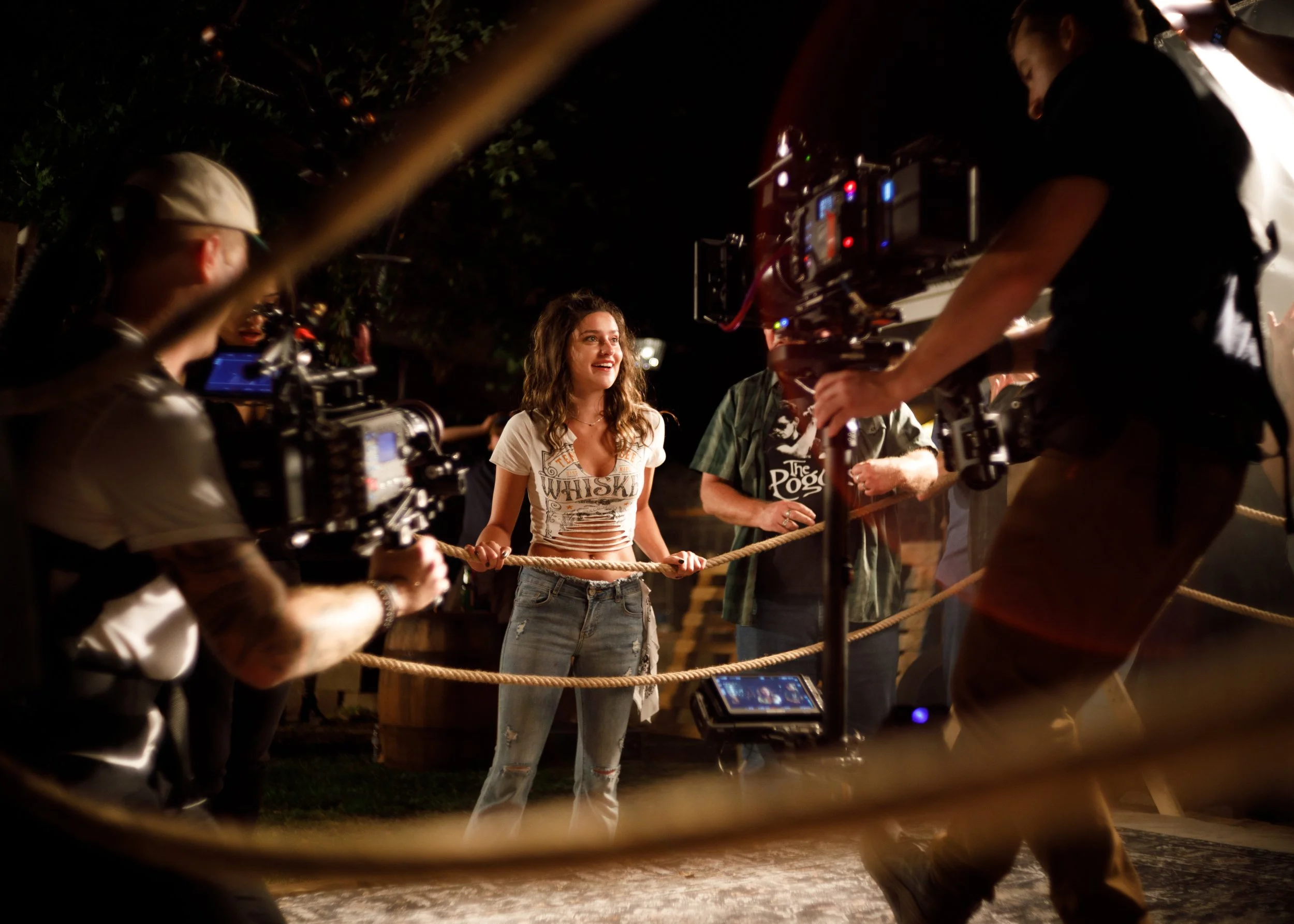 A woman with curly hair and a white t-shirt standing behind a rope barrier while cameras and crew filming her at night outdoors.