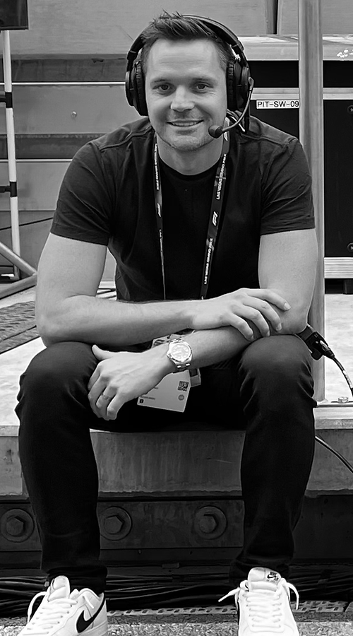A young man with short hair and light skin, wearing a headset with a microphone, a black T-shirt, black pants, and white sneakers, sitting and smiling at the camera in an indoor setting with shelves and equipment in the background.