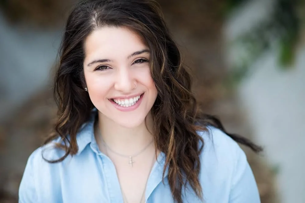 A woman with long wavy brown hair and a nose piercing smiling in an outdoor setting.