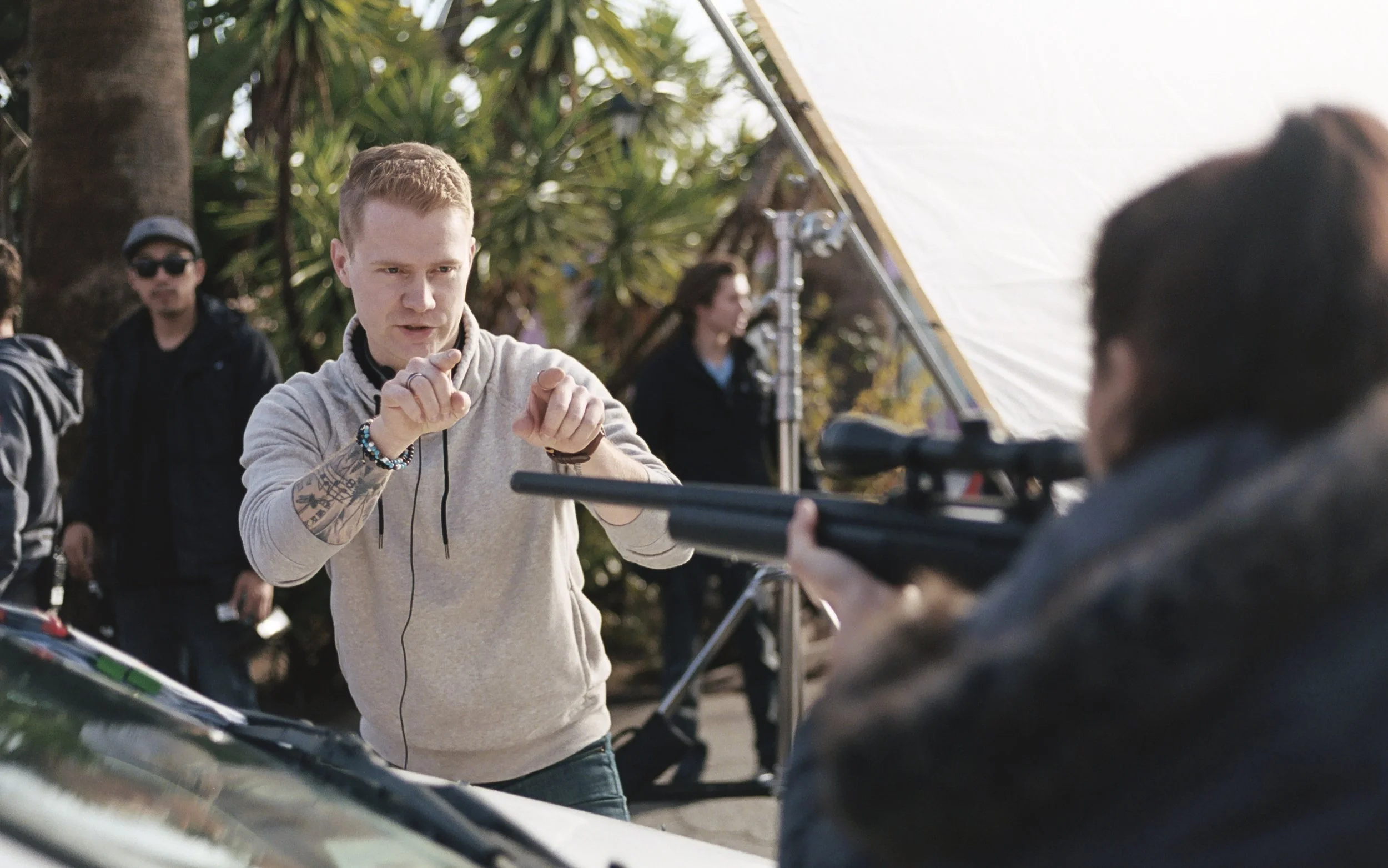 A man with tattoos on his arms is holding a clapboard while filming a scene outdoors. Several people and trees are visible in the background.