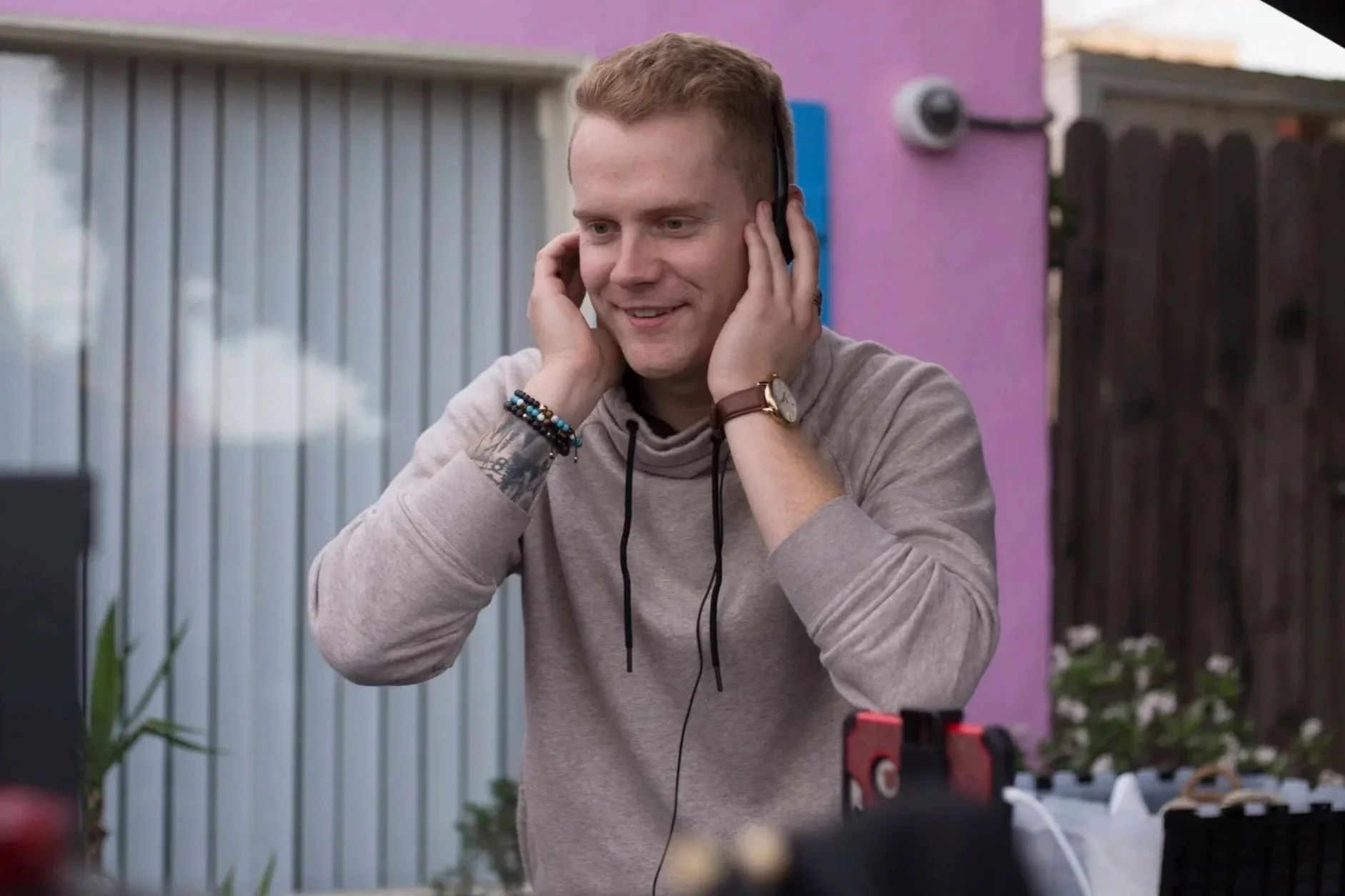 A young man with short red hair, wearing a light gray hoodie and a watch, smiling while listening to headphones outdoors in a backyard or patio area.