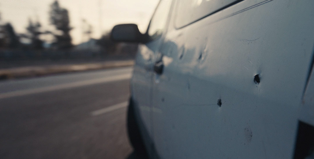 Close-up of a white car with bullet holes on the side door, driving on a highway.