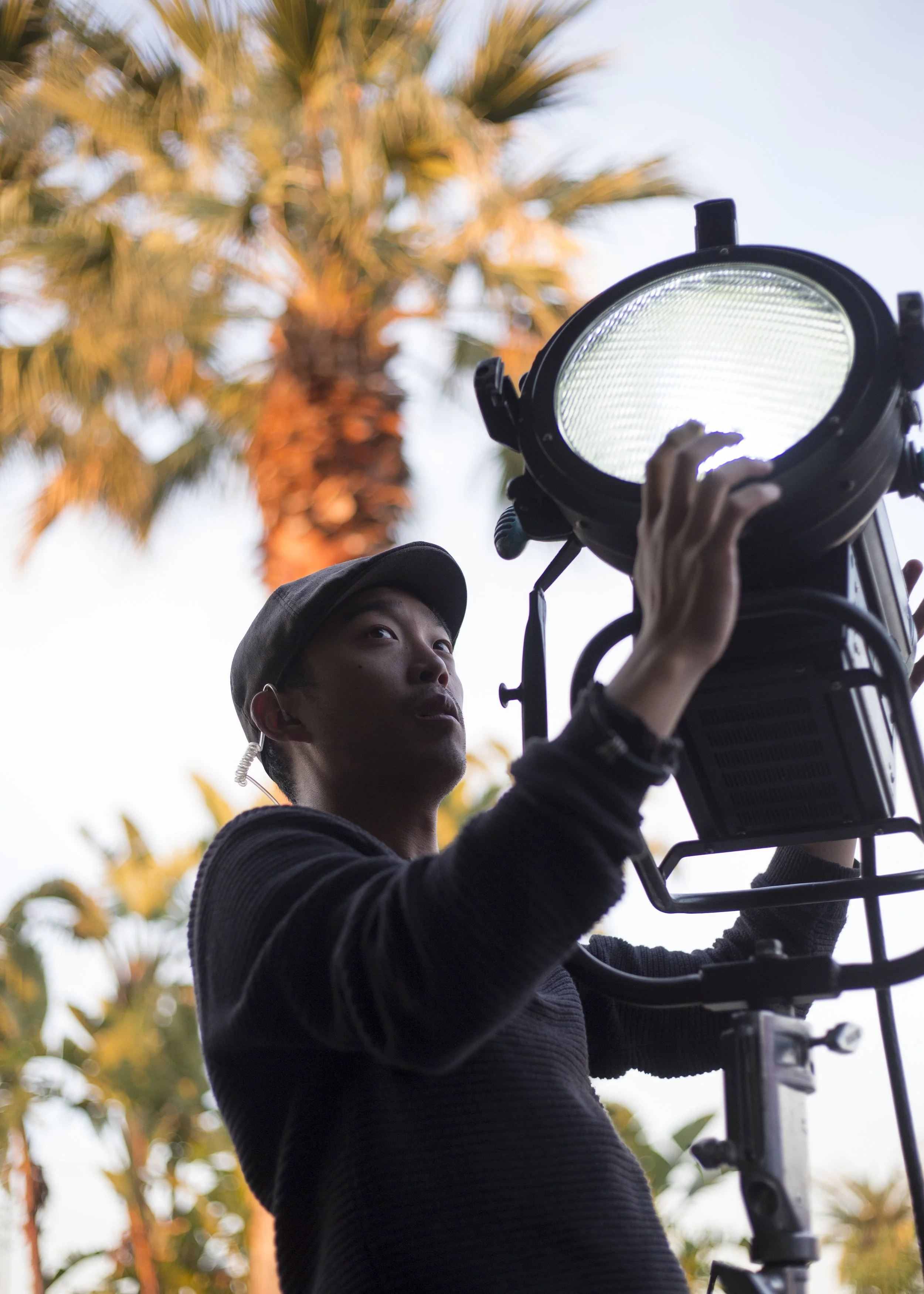 A person wearing a black hoodie and a cap operating a large film or photography light outdoors, with palm trees and a bright sky in the background.