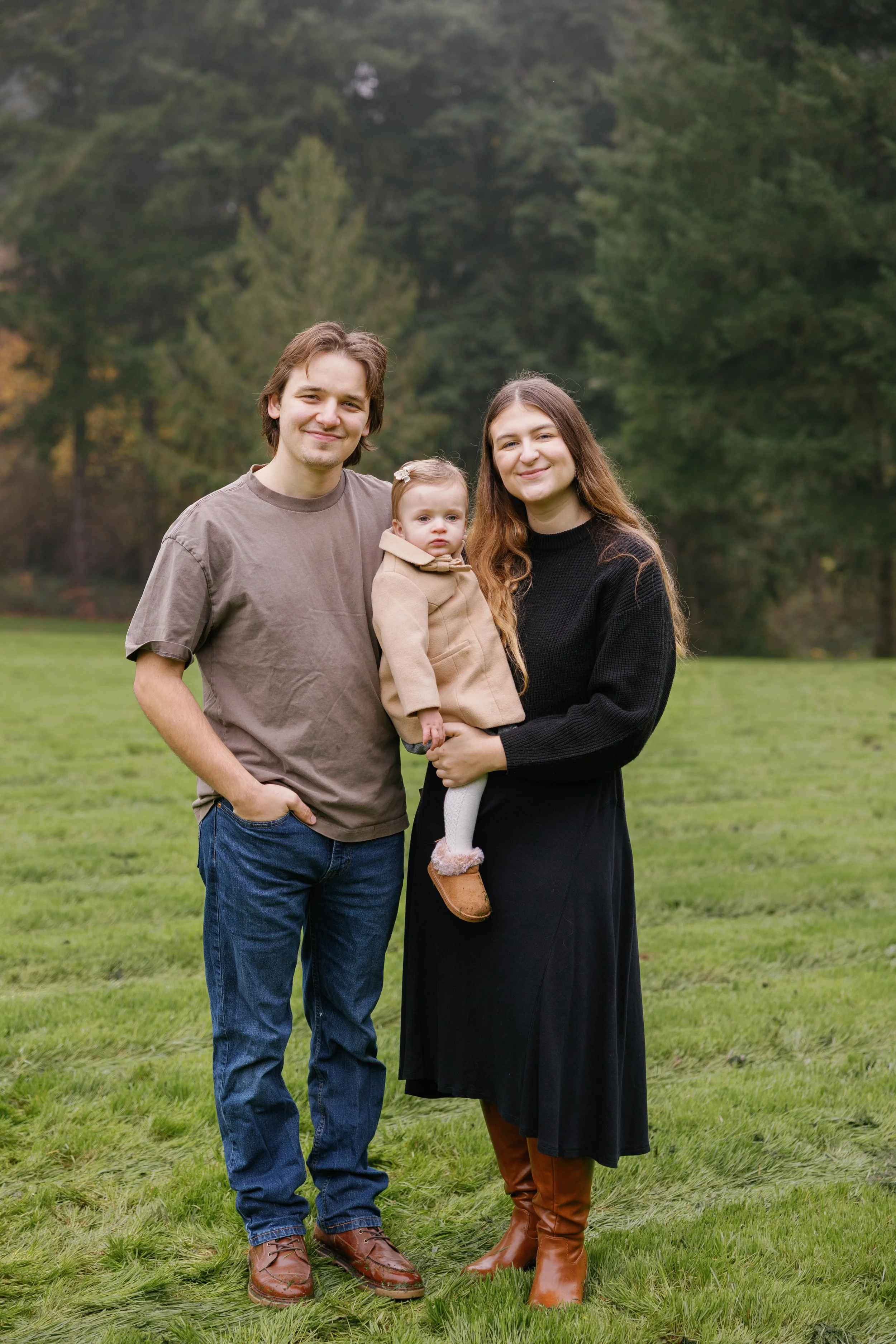 A family of three standing outdoors on grass with trees in the background. An adult man with brown hair wearing a gray t-shirt and jeans, an adult woman with long brown hair wearing a black sweater and a black skirt, and a young girl with light brown hair in a beige coat, brown boots, and a bow in her hair.
