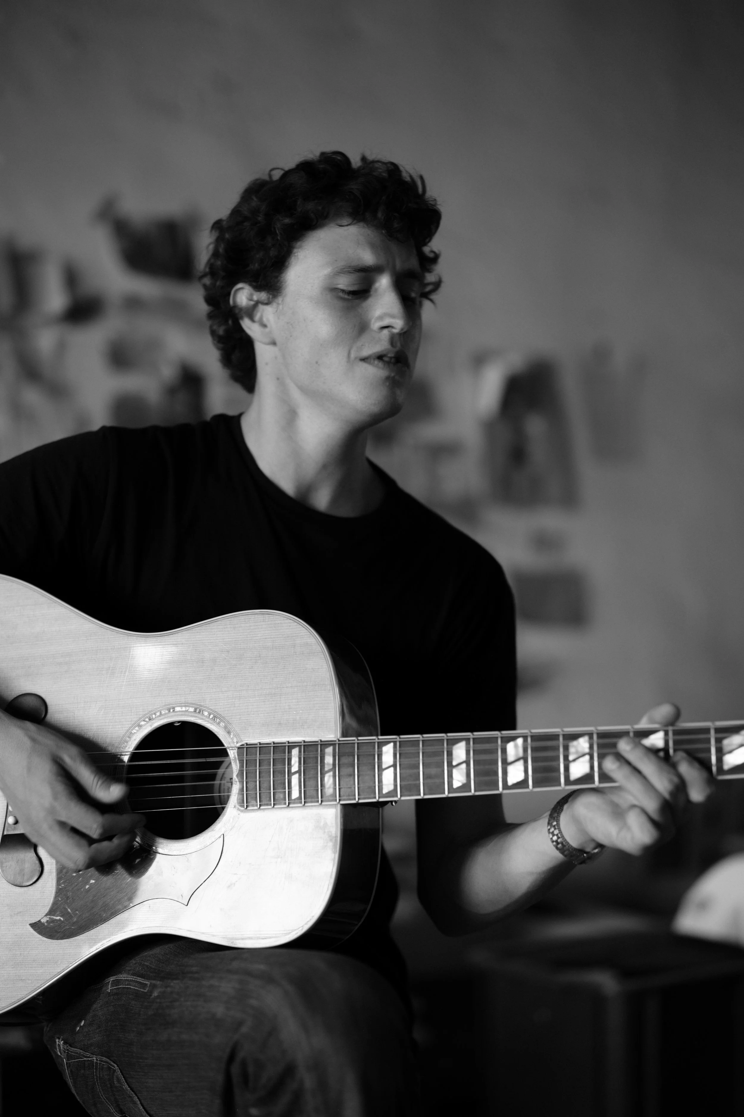 A young man with curly hair playing an acoustic guitar, wearing a black t-shirt, sitting indoors with a blurred background.