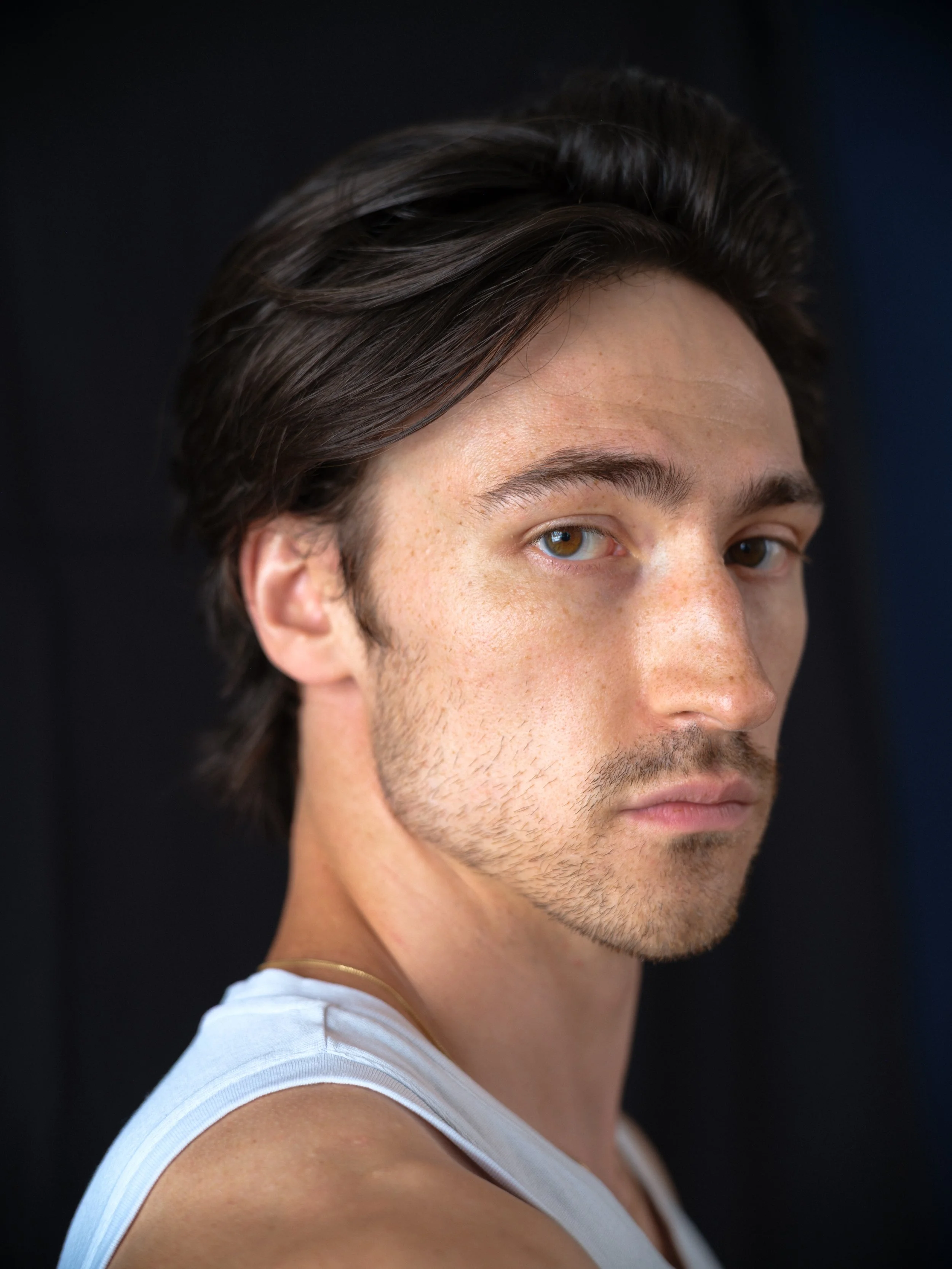 Close-up of a young man's face, showing brown hair, hazel eyes, fair skin with freckles, and part of a white tank top against a dark background.