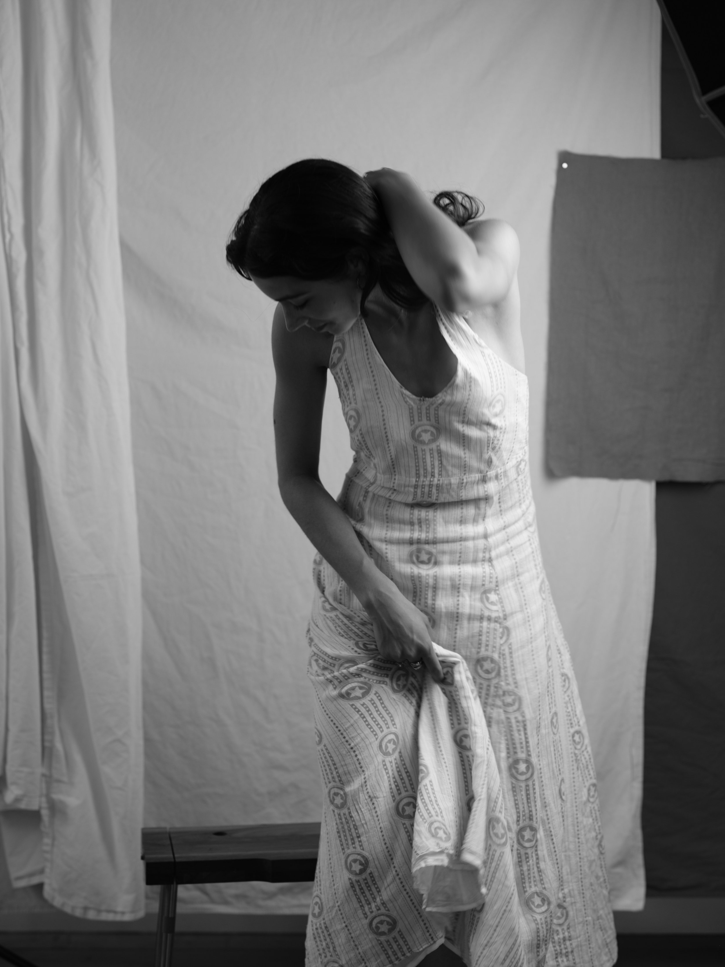 A woman with long dark hair wearing a patterned dress, bending down to pick up a piece of cloth in a photography studio with a plain backdrop and fabric curtains.
