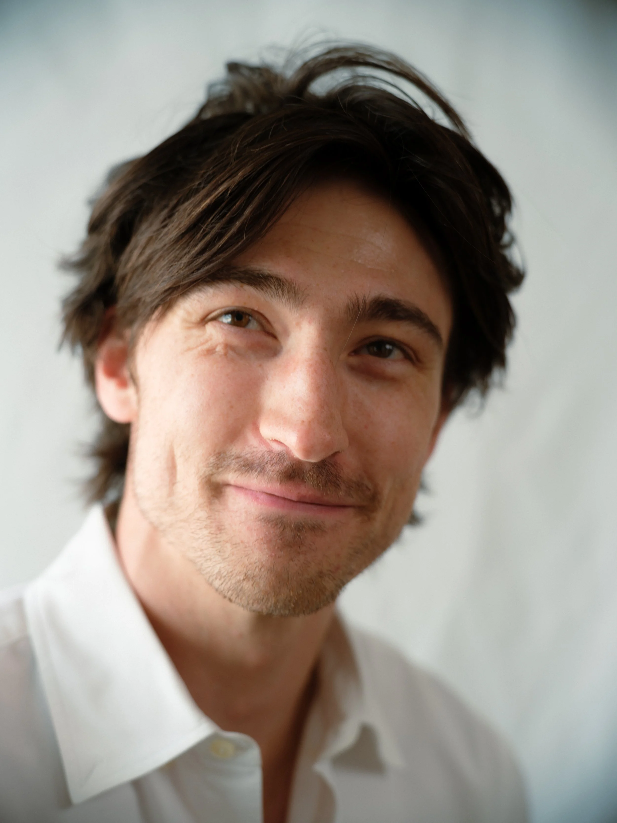 Close-up of a smiling man with dark hair, wearing a white shirt, looking at the camera against a plain background.