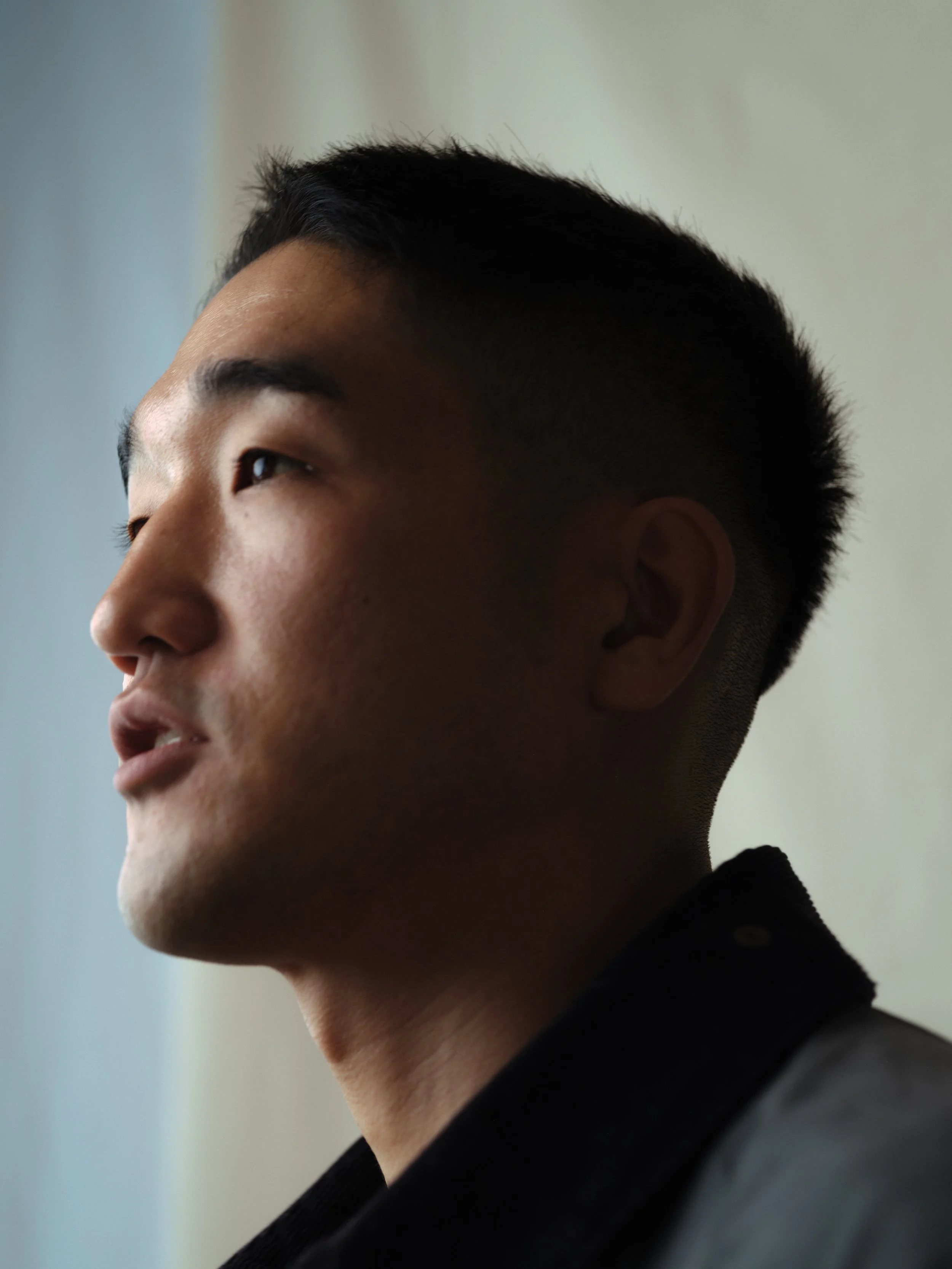 Close-up side profile of a young man with short black hair, wearing a dark top, with a blurred background.