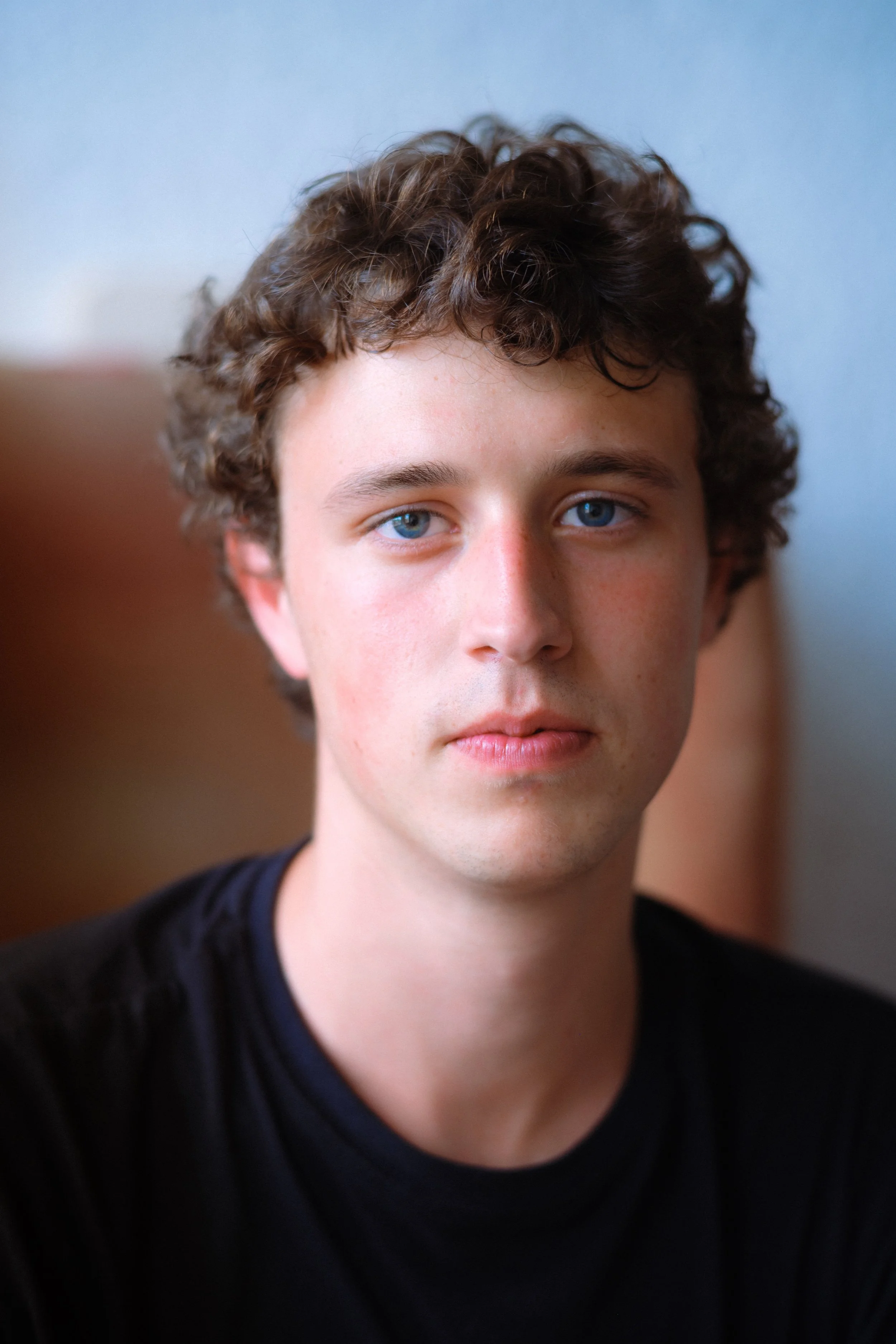 A young man with curly brown hair and blue eyes taking a selfie indoors, wearing a black shirt.