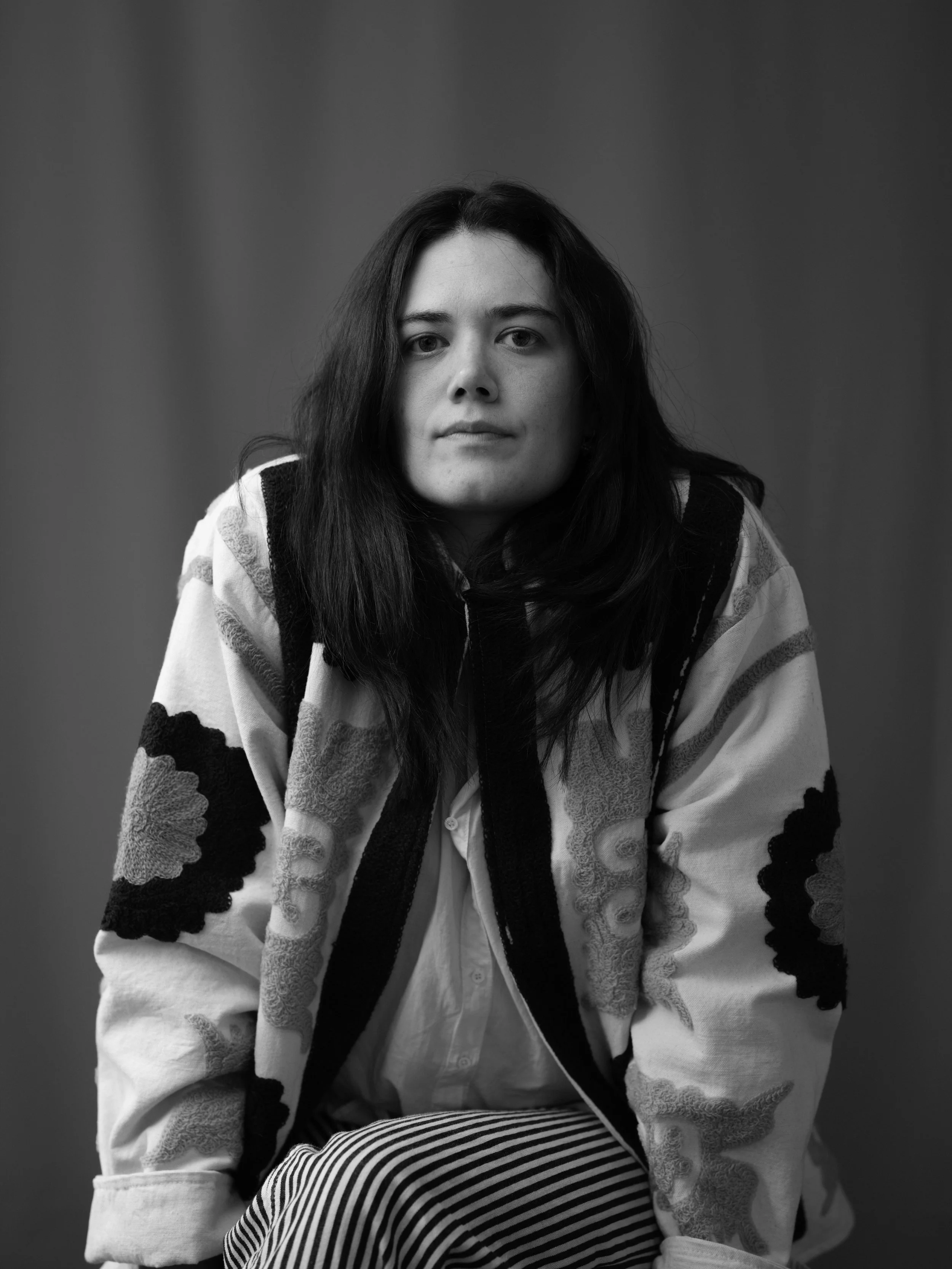 Black and white photo of a young woman with long dark hair, sitting indoors against a plain background. She is wearing a patterned hoodie over a button-up shirt, and striped pants, looking directly at the camera with a neutral expression.
