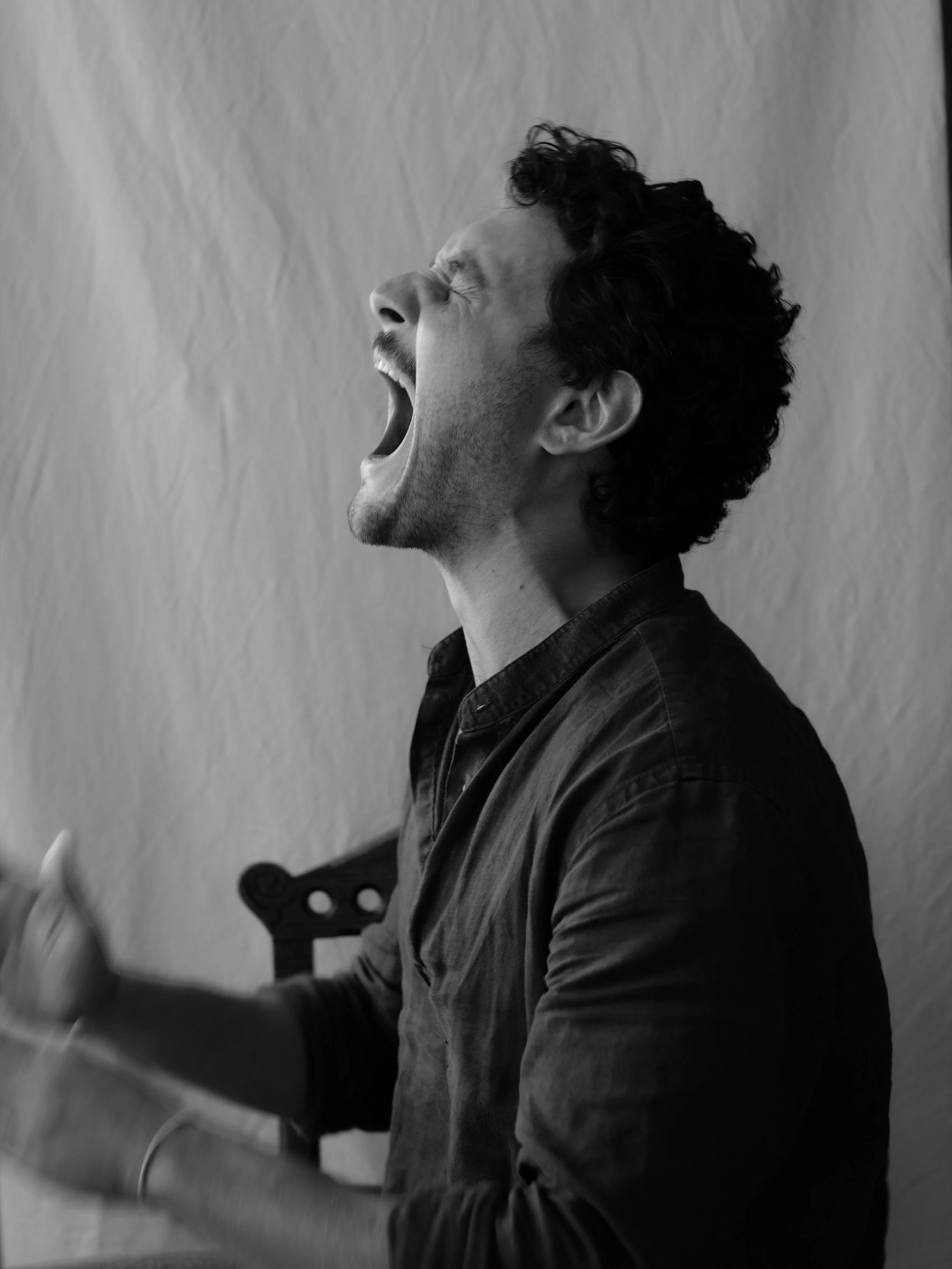 Black and white photo of a man with curly hair opening his mouth wide and eyes closed, appearing to yawn or shout.