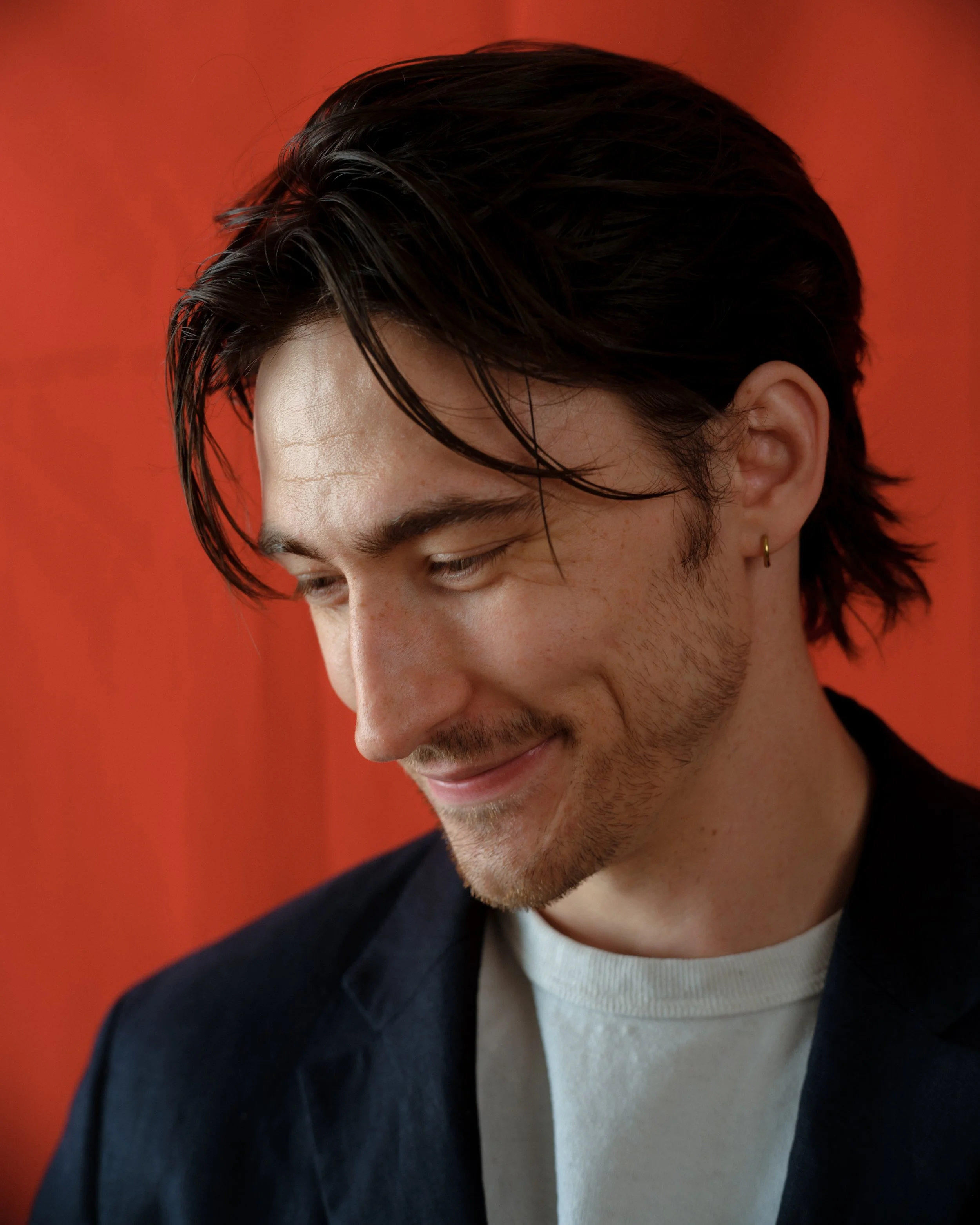 Close-up of a young man with brown hair, styled slightly messy, wearing a gold earring, black blazer, white t-shirt, and a faint smile, against a red background.
