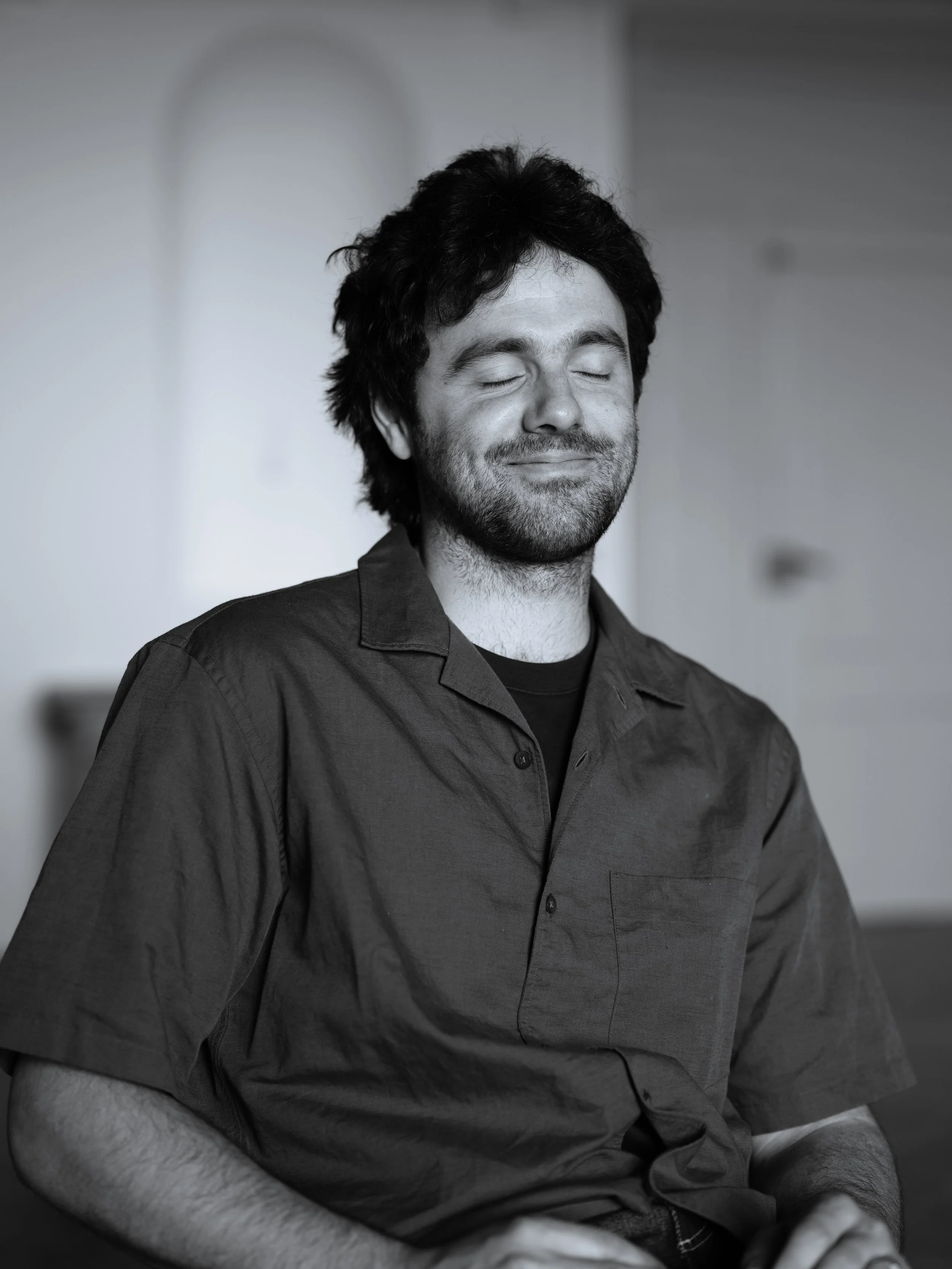 A man with closed eyes, dark wavy hair, and facial hair, wearing a dark collared shirt, sitting and smiling softly in a relaxed pose