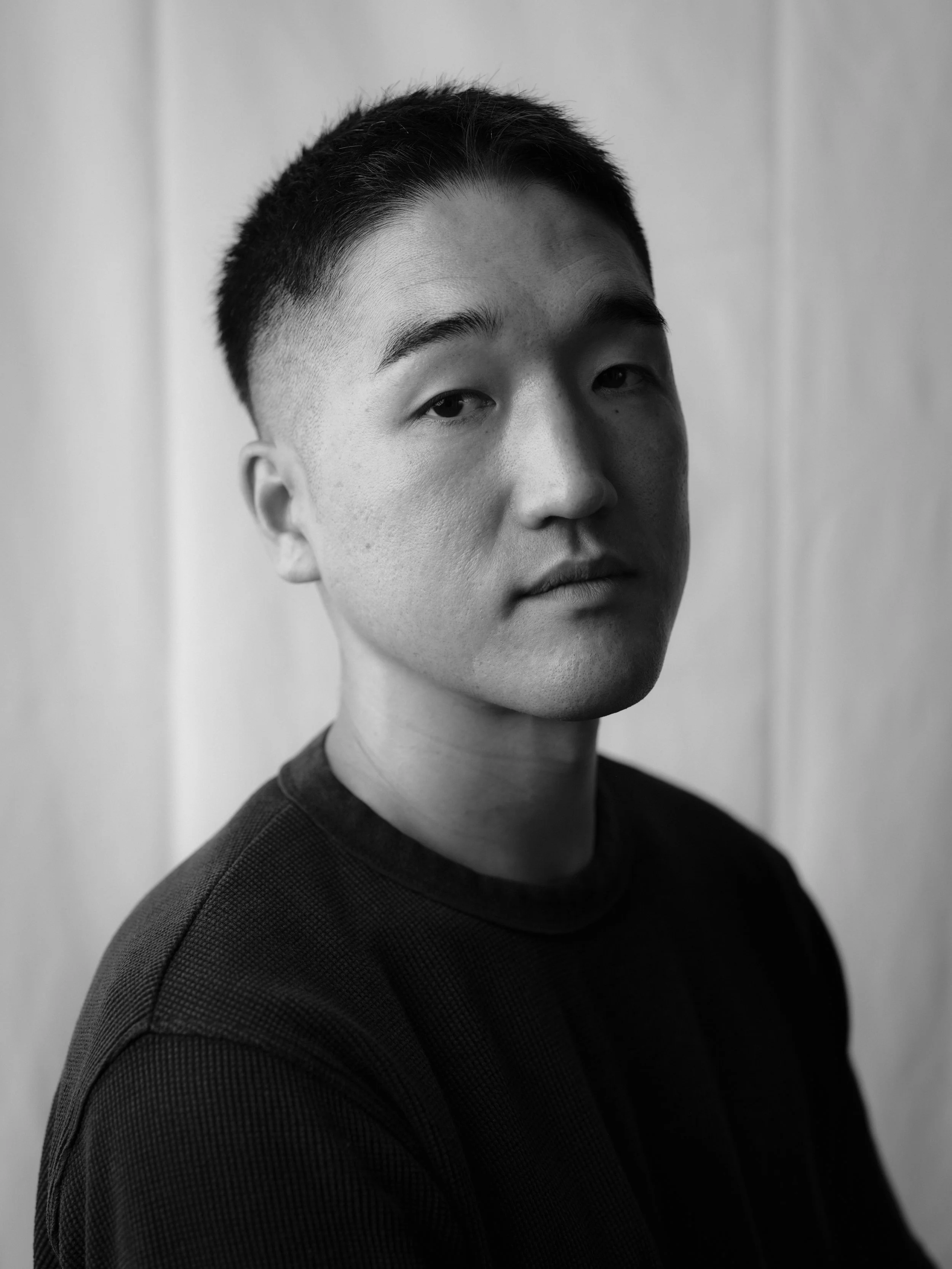 Black and white portrait of a young man with short hair, wearing a dark textured shirt, against a plain background.