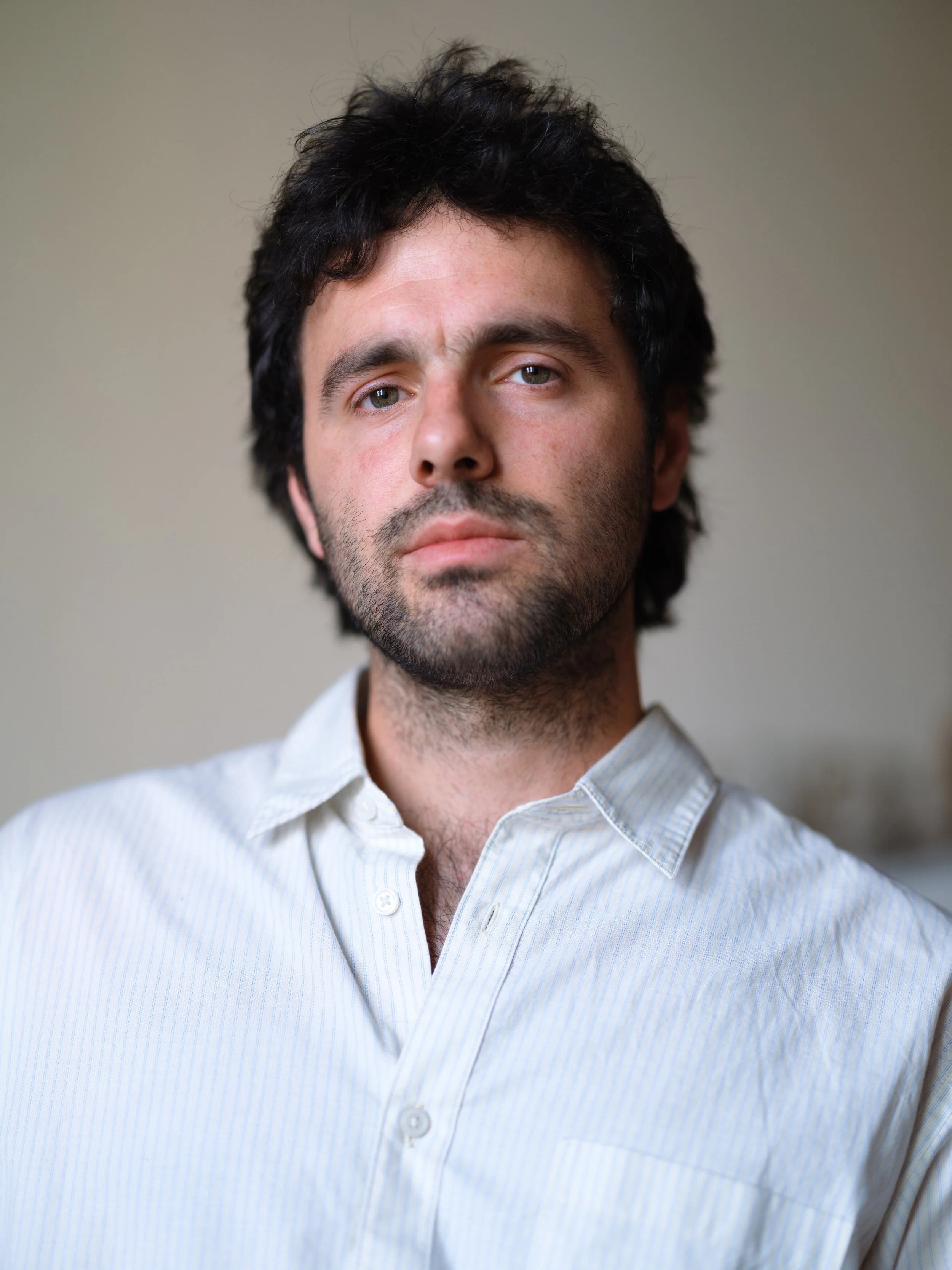 A man with dark, curly hair, light skin, and blue eyes wearing a light blue, striped button-up shirt.