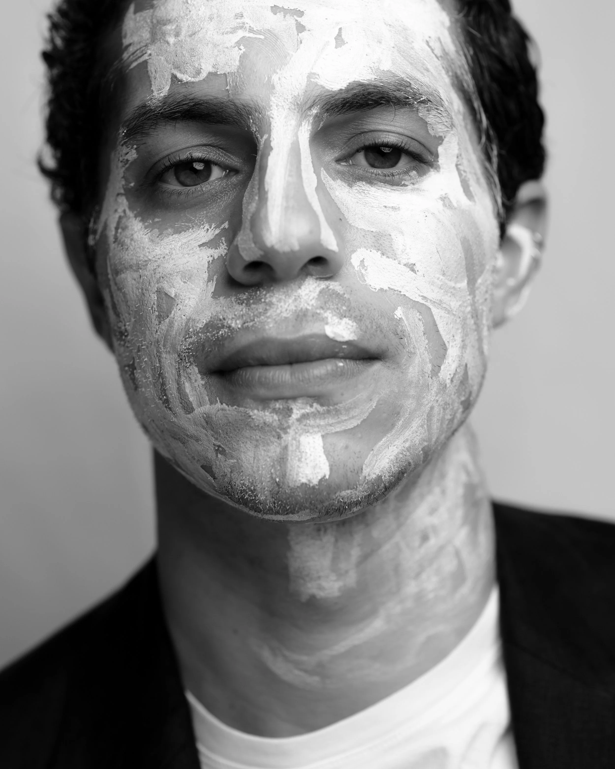 Close-up black-and-white photo of a young man with short dark hair and light skin, with a white paint or plaster on his face, covering parts of his forehead, nose, cheeks, and chin.