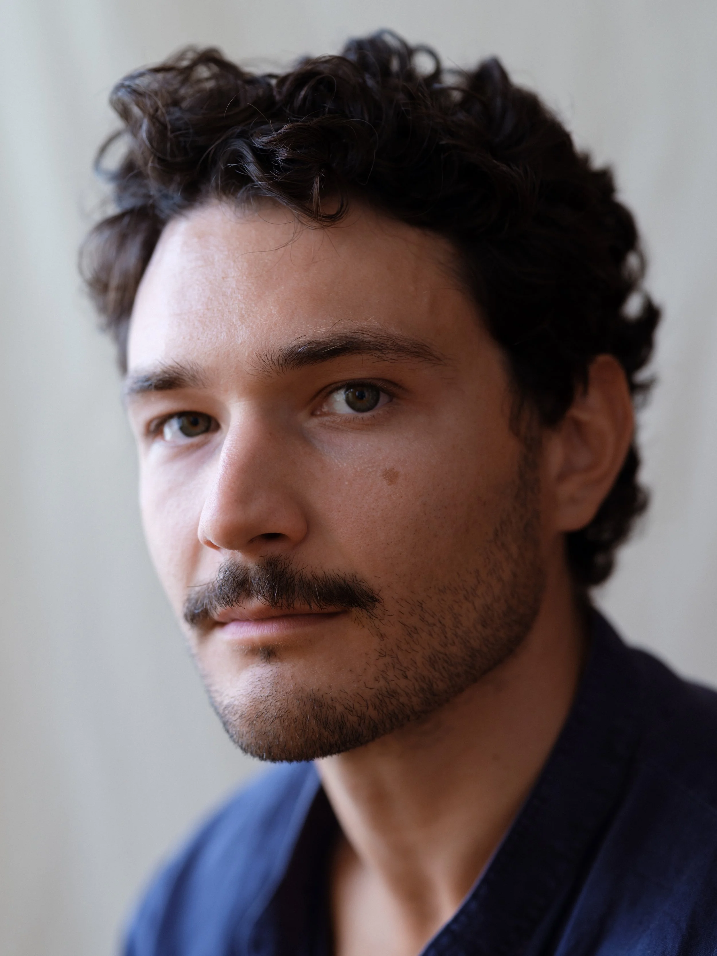 Close-up of a young man with dark curly hair, light skin, and a light beard, wearing a dark jacket over a blue shirt, looking slightly to the side.