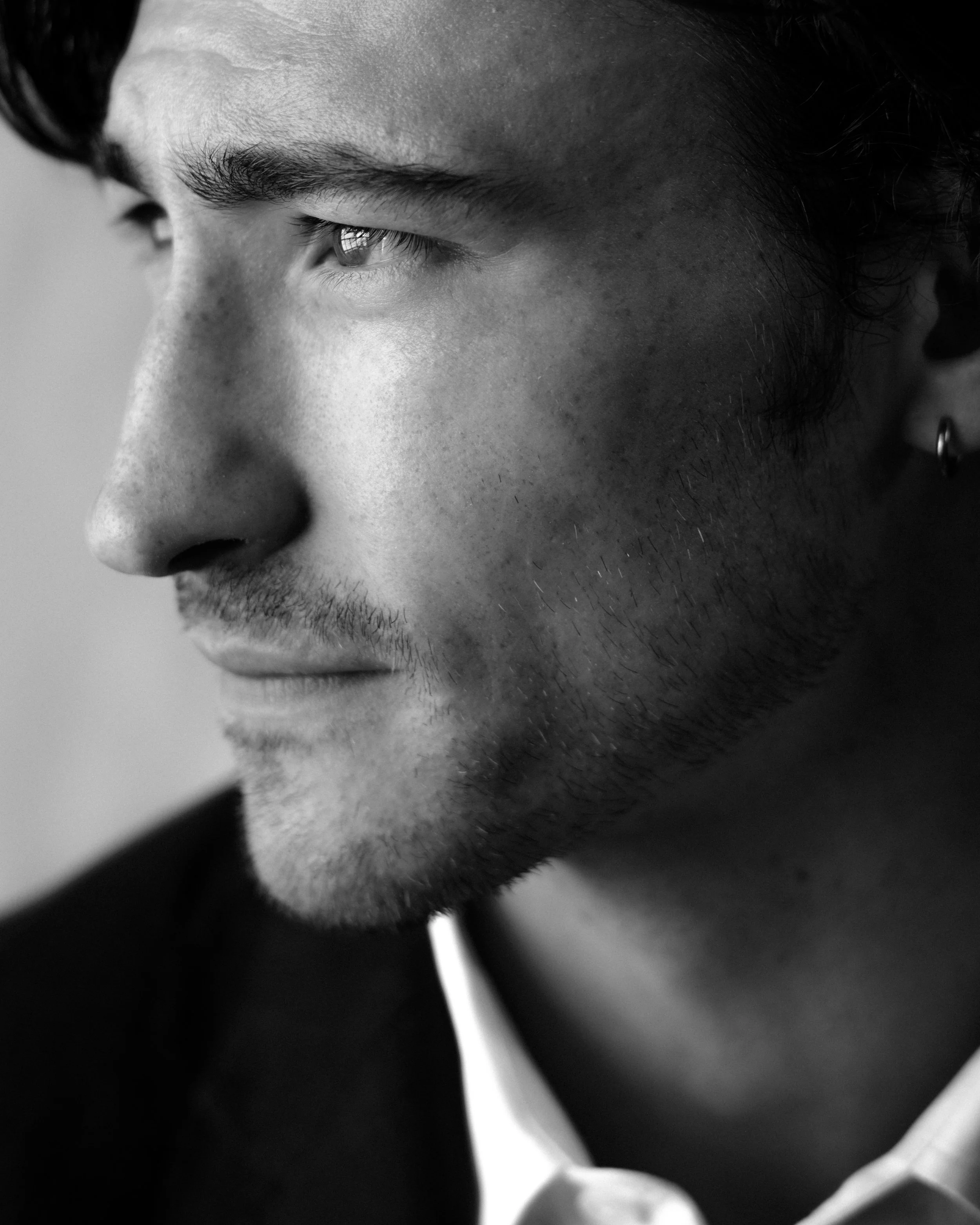 Close-up of a young man's face in black and white, showing his eye, nose, and part of his lips and chin, with dark hair and an earring visible on the ear.