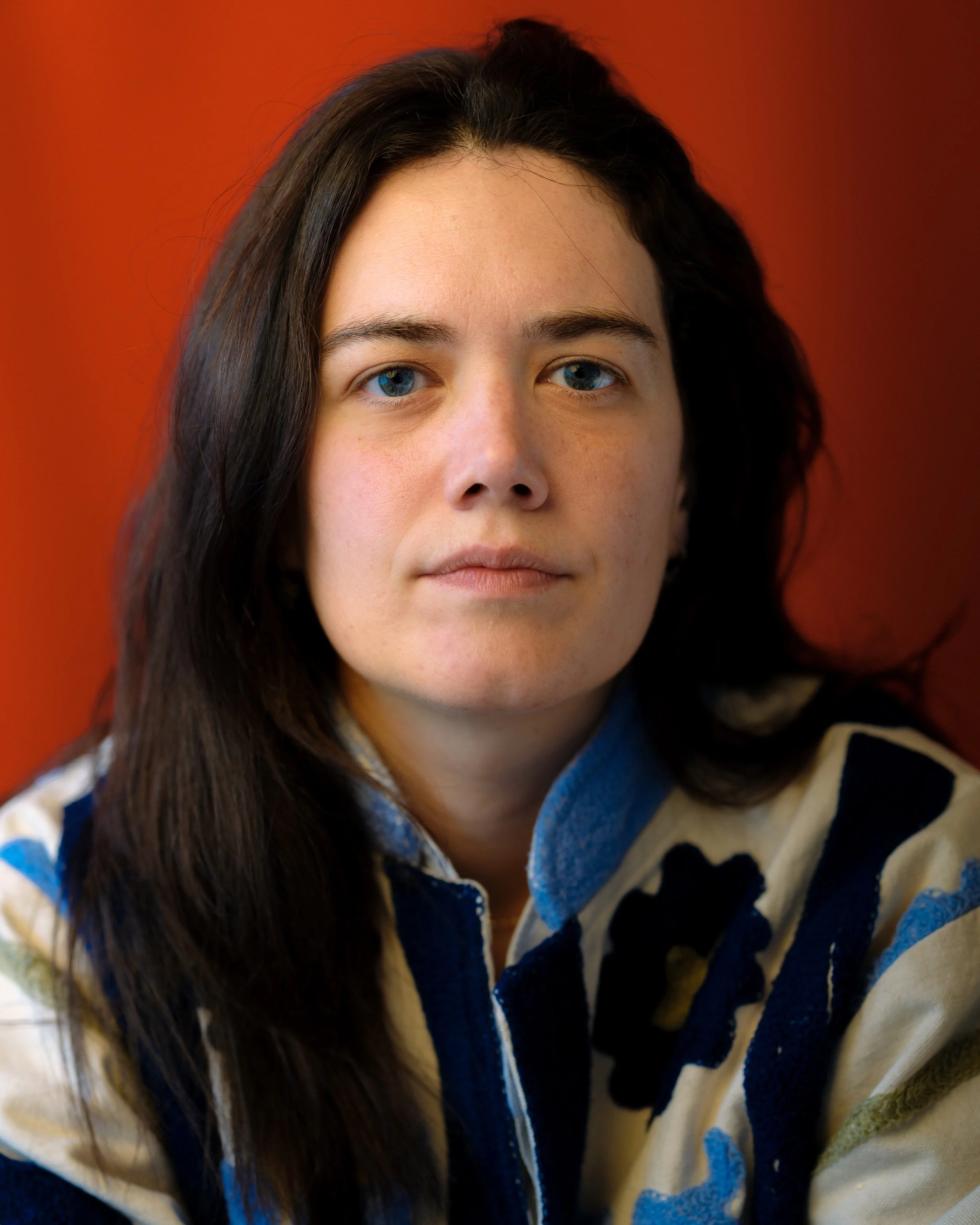 Close-up of a woman with long dark hair and blue eyes, wearing a cream and navy blue patterned fleece jacket, against an orange background.