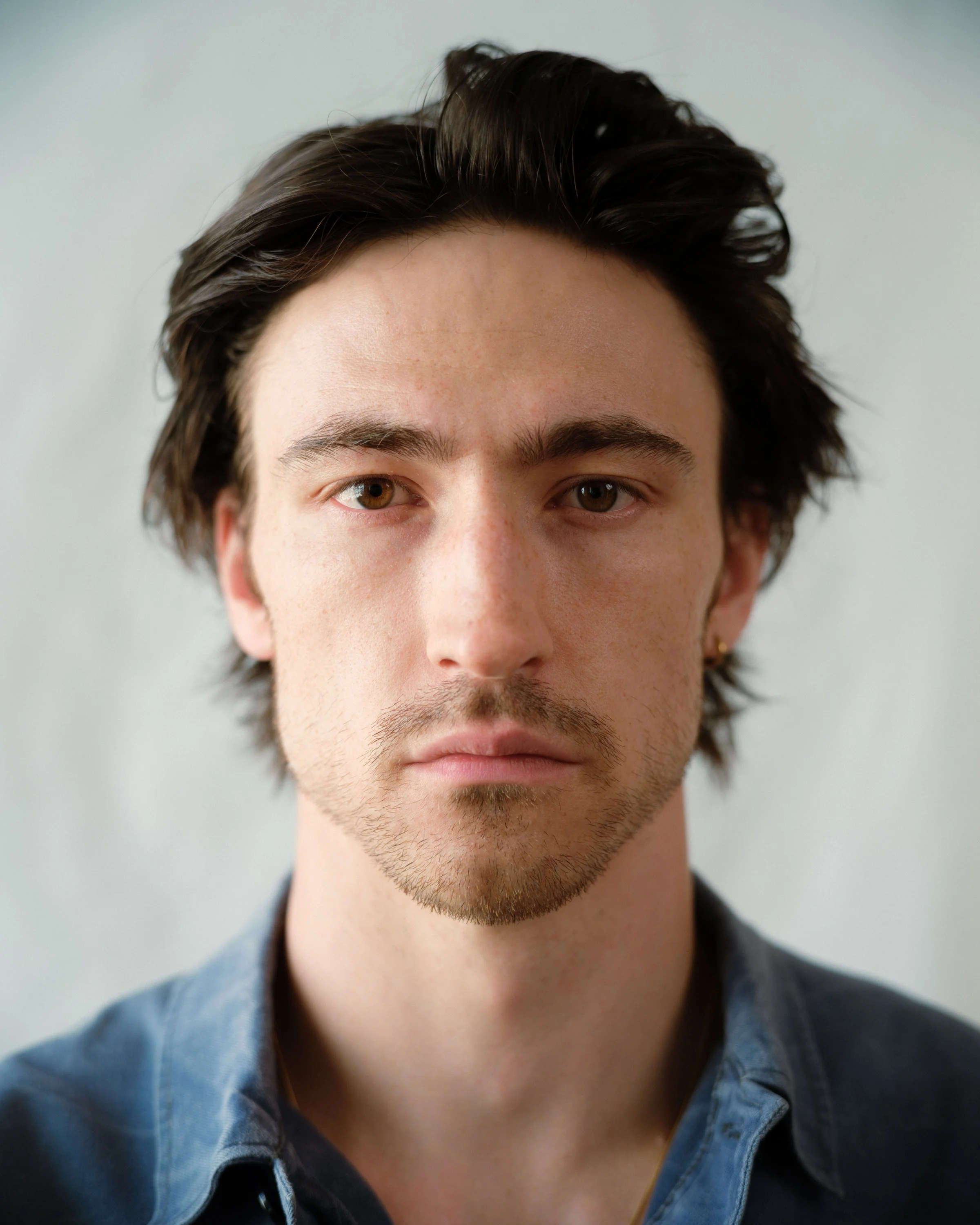 Close-up of a young man with brown hair, brown eyes, light skin, wearing a denim shirt, looking directly at camera against a neutral background.