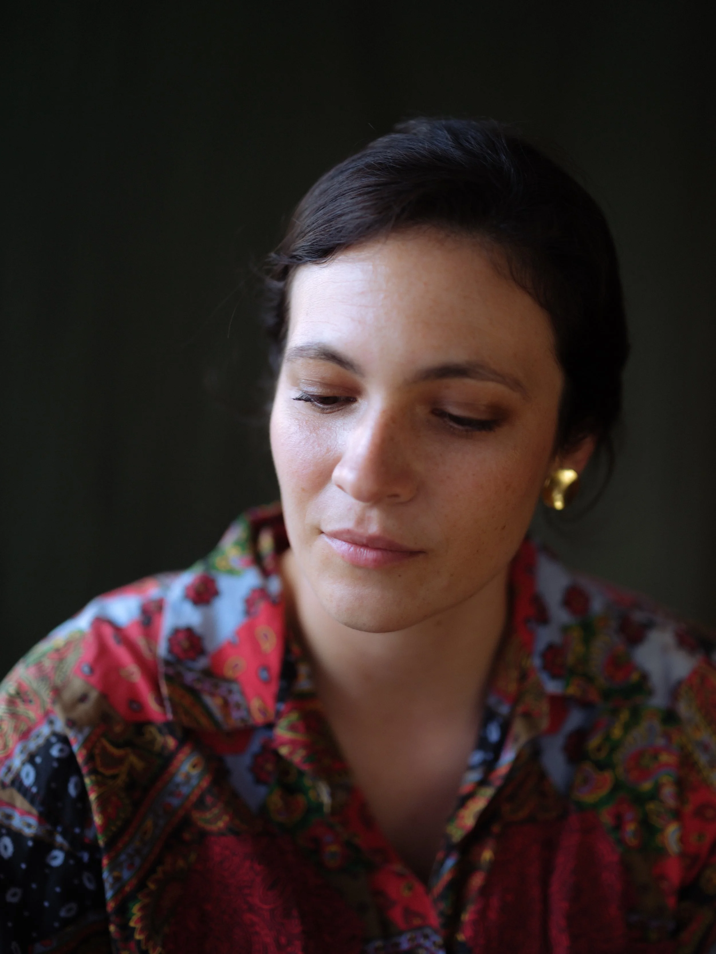 Close-up of a woman with dark hair and gold earrings, wearing a colorful, patterned shirt, looking downward with a neutral expression.