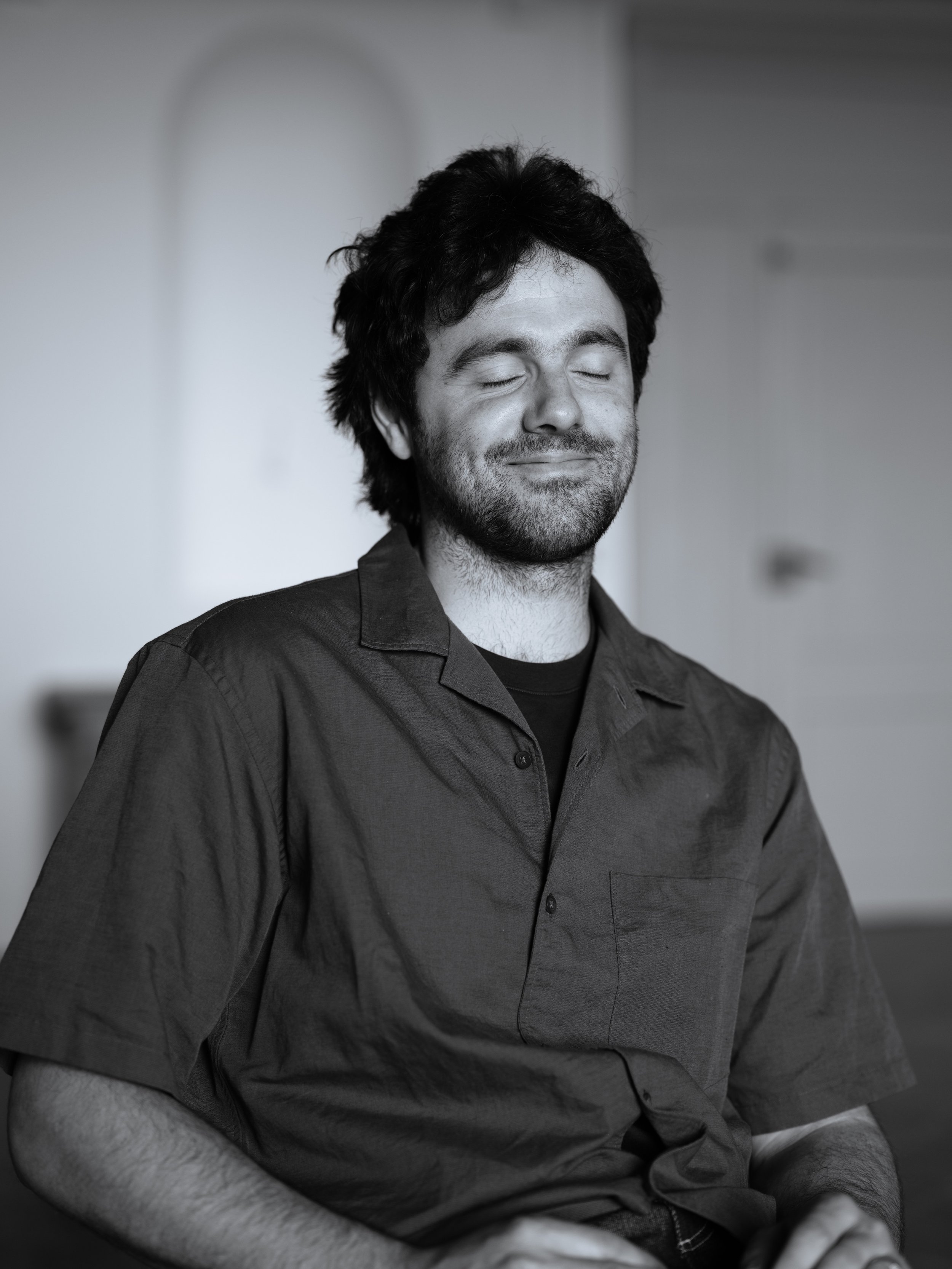Black and white photo of a smiling man with closed eyes, wearing a button-up shirt, sitting with his hands folded, in a relaxed, peaceful pose.