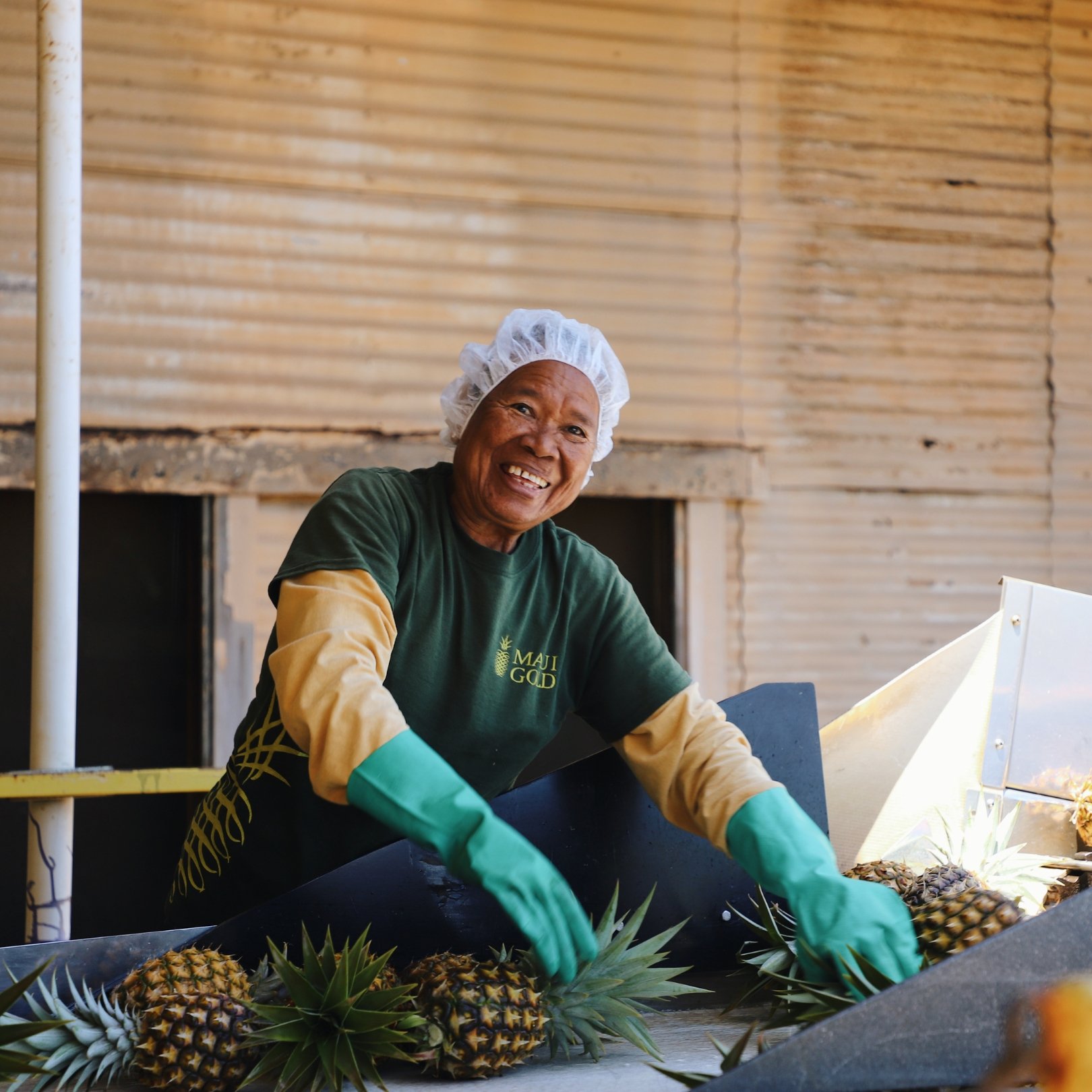 A smiling woman in green gloves and hairnet handling pineapples at a stall in a market or food processing area with a wooden wall background.