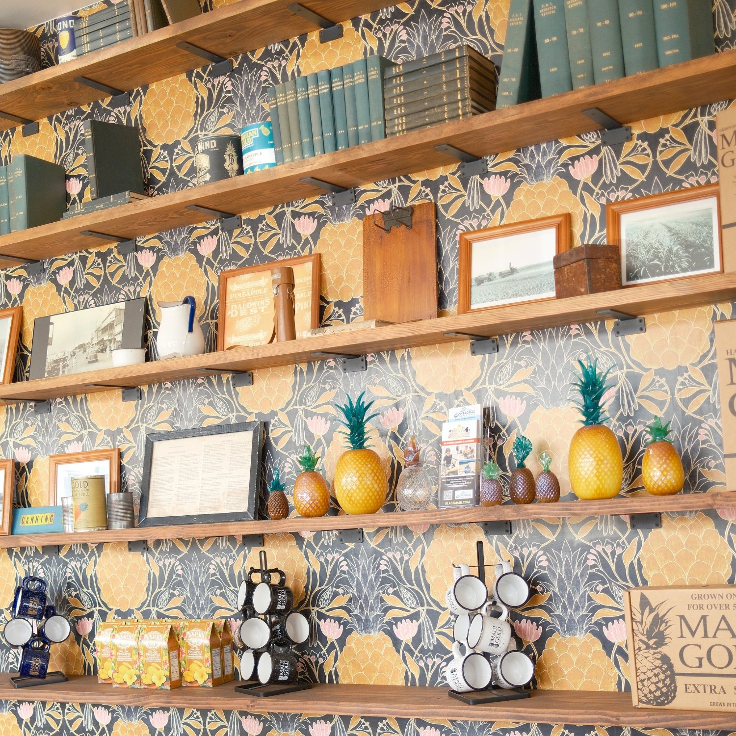 Wooden shelves with books, picture frames, pineapple-shaped decorations, and coffee cups against a floral wallpaper background.