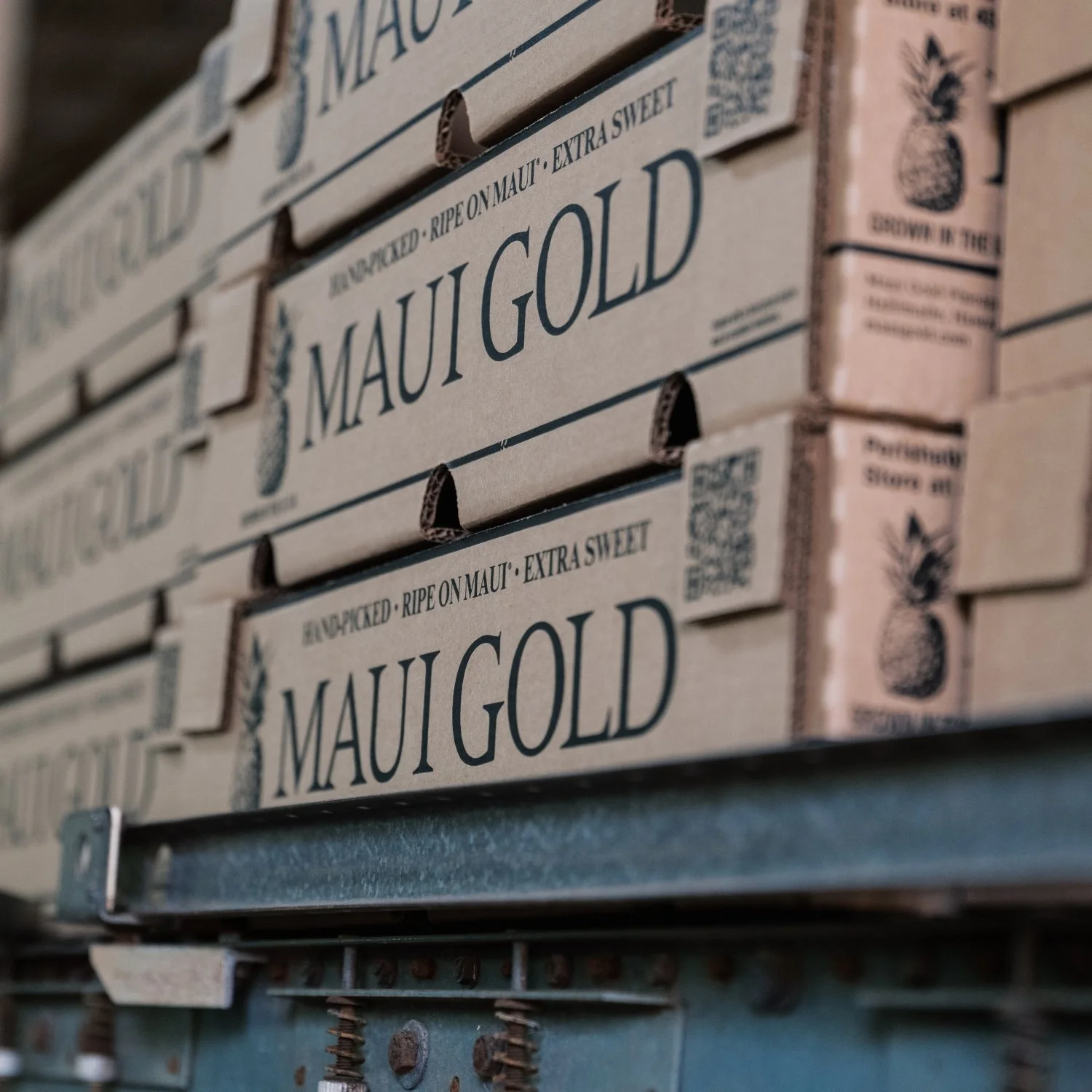 Close-up of stacked cardboard boxes labeled 'Maui Gold,' indicating they contain ripe, extra sweet pineapple from Maui with company branding and QR codes.