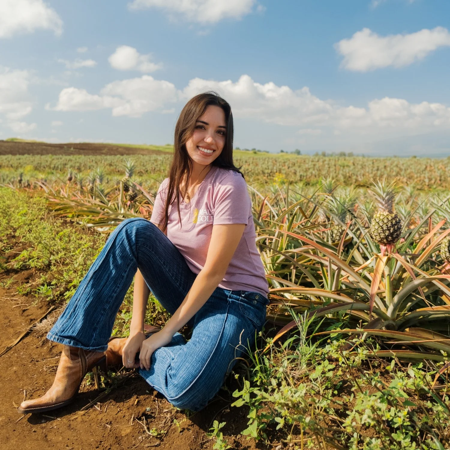 A young woman sitting on the ground in a pineapple field on a sunny day, smiling at the camera with a background of pineapple plants and a blue sky with clouds.