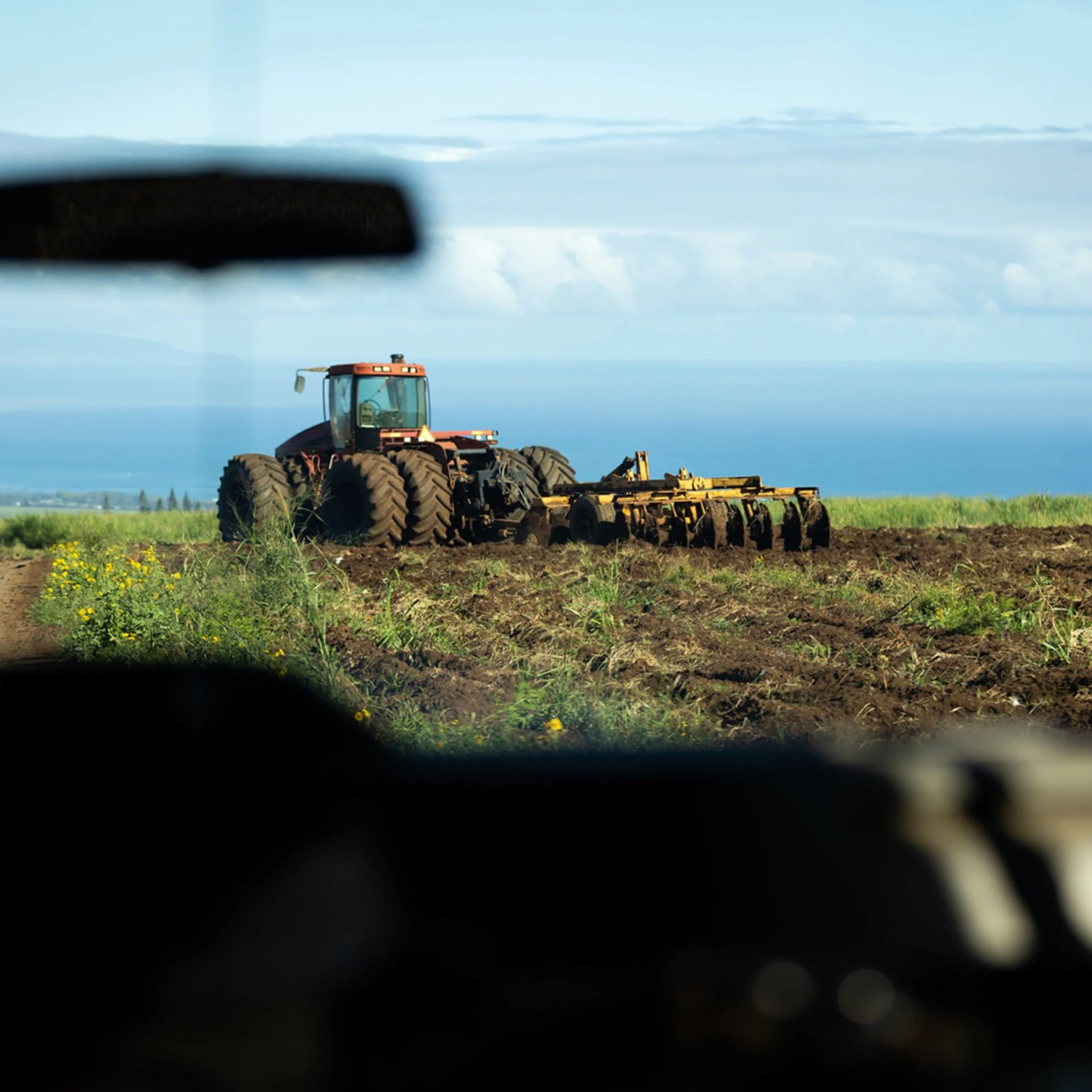 A tractor working in a field during daytime, seen through the window of a vehicle, with a cloudy sky overhead.
