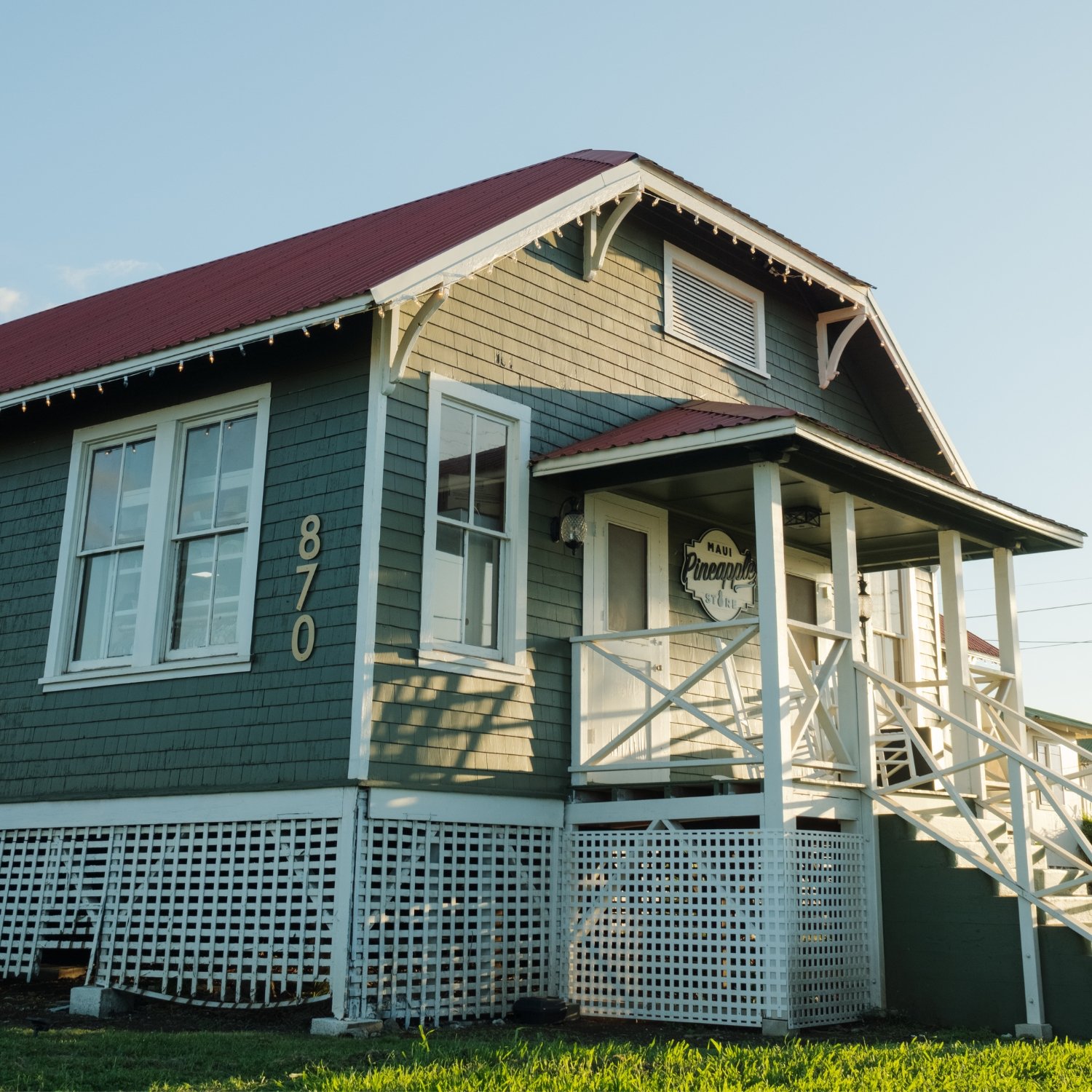 A two-story house painted in green with white trim, featuring a front porch with white railings and stairs, a sign reading "Maui Pineapple Store," and a fence at the bottom. The house has multiple windows, a gabled roof with red shingles, and a clear
