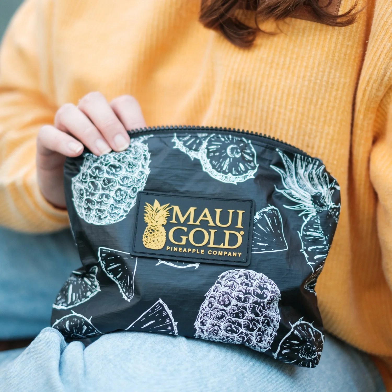 A person in a yellow sweater is holding a black Maui Gold Pineapple Company snack bag with pineapple and fruit illustrations.