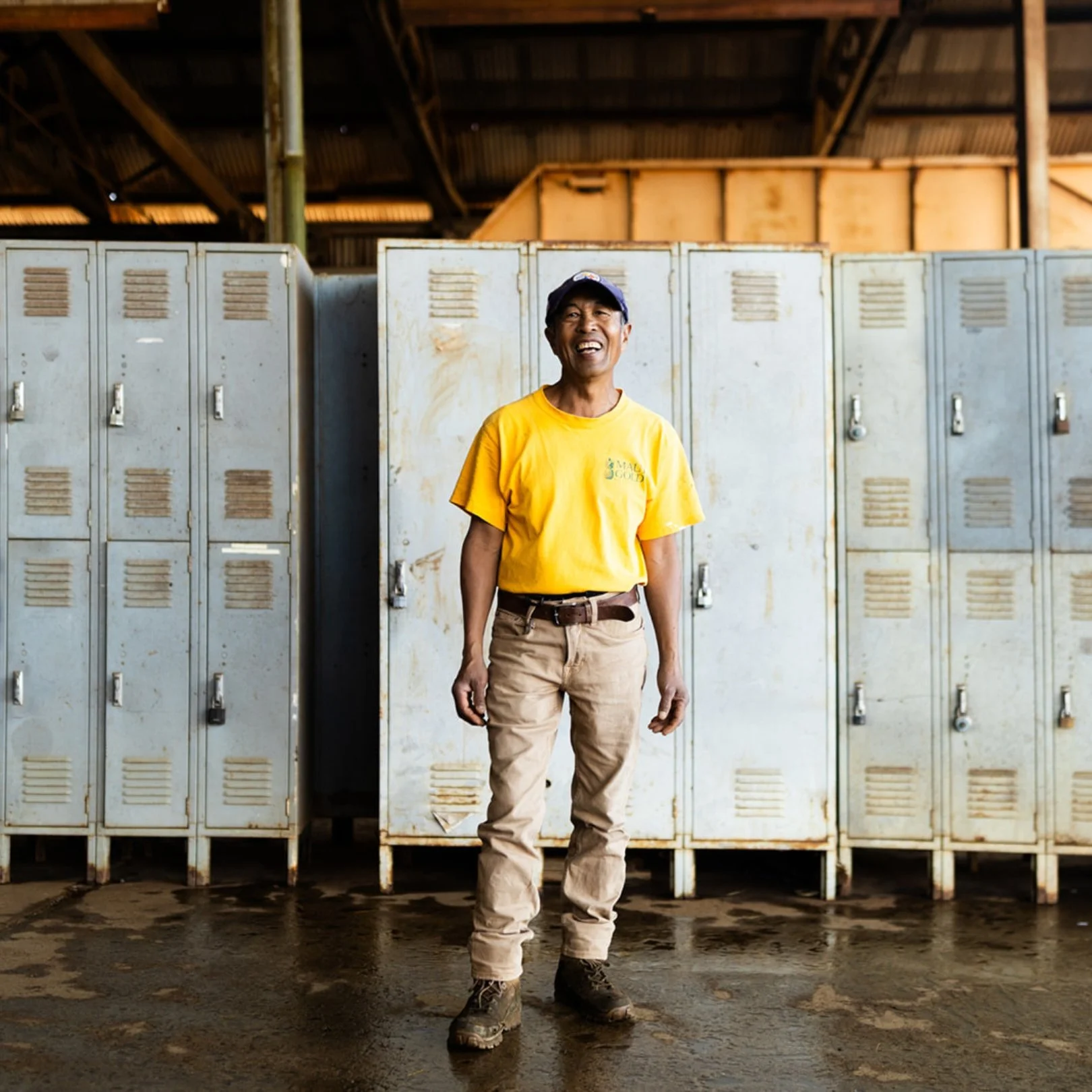 A man wearing a yellow T-shirt, khaki pants, and work boots, standing in front of a row of white lockers inside a rustic warehouse or storage facility with a metal roof.