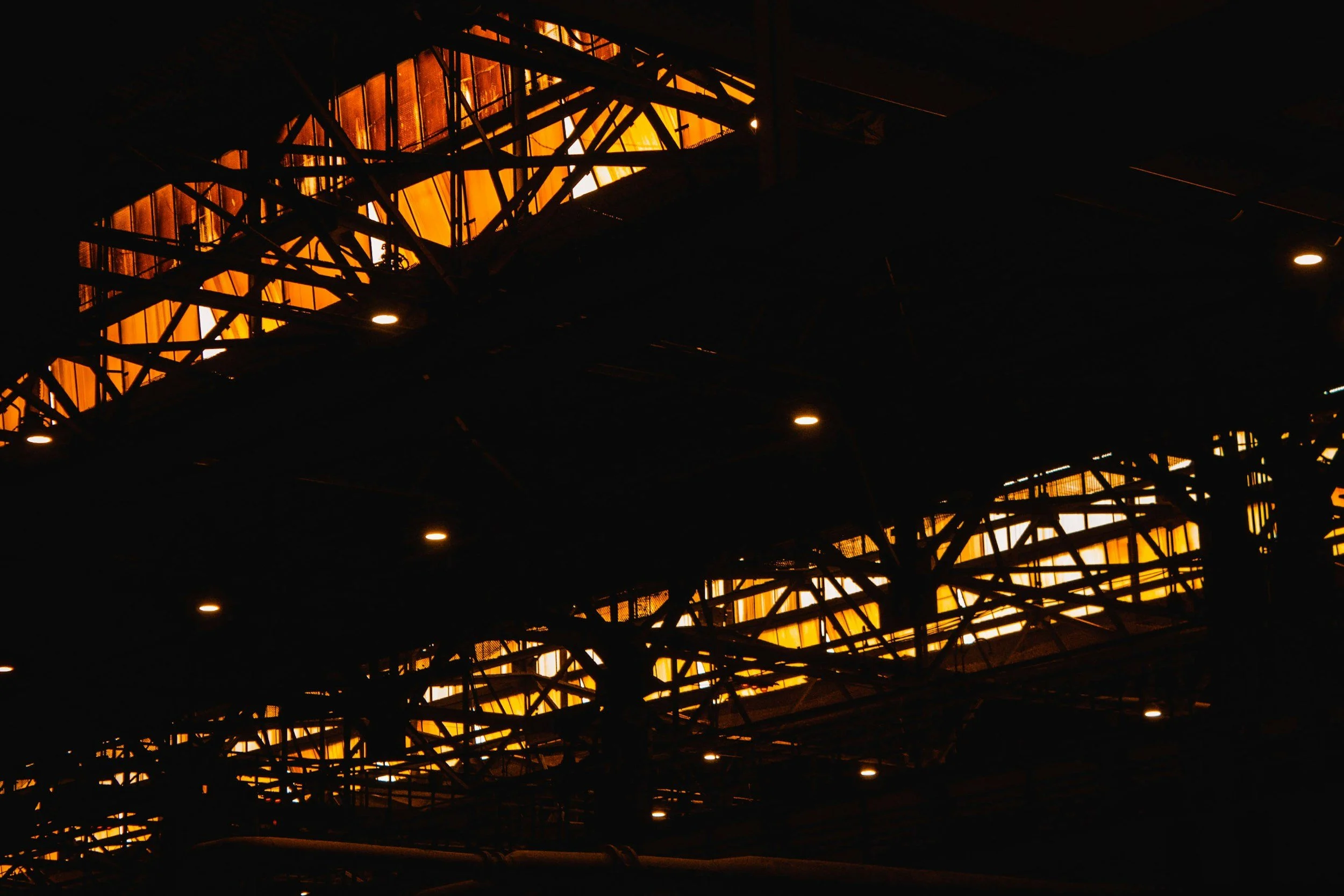 View of a large industrial or architectural structure with illuminated orange panels seen through black steel framework at dusk or night.
