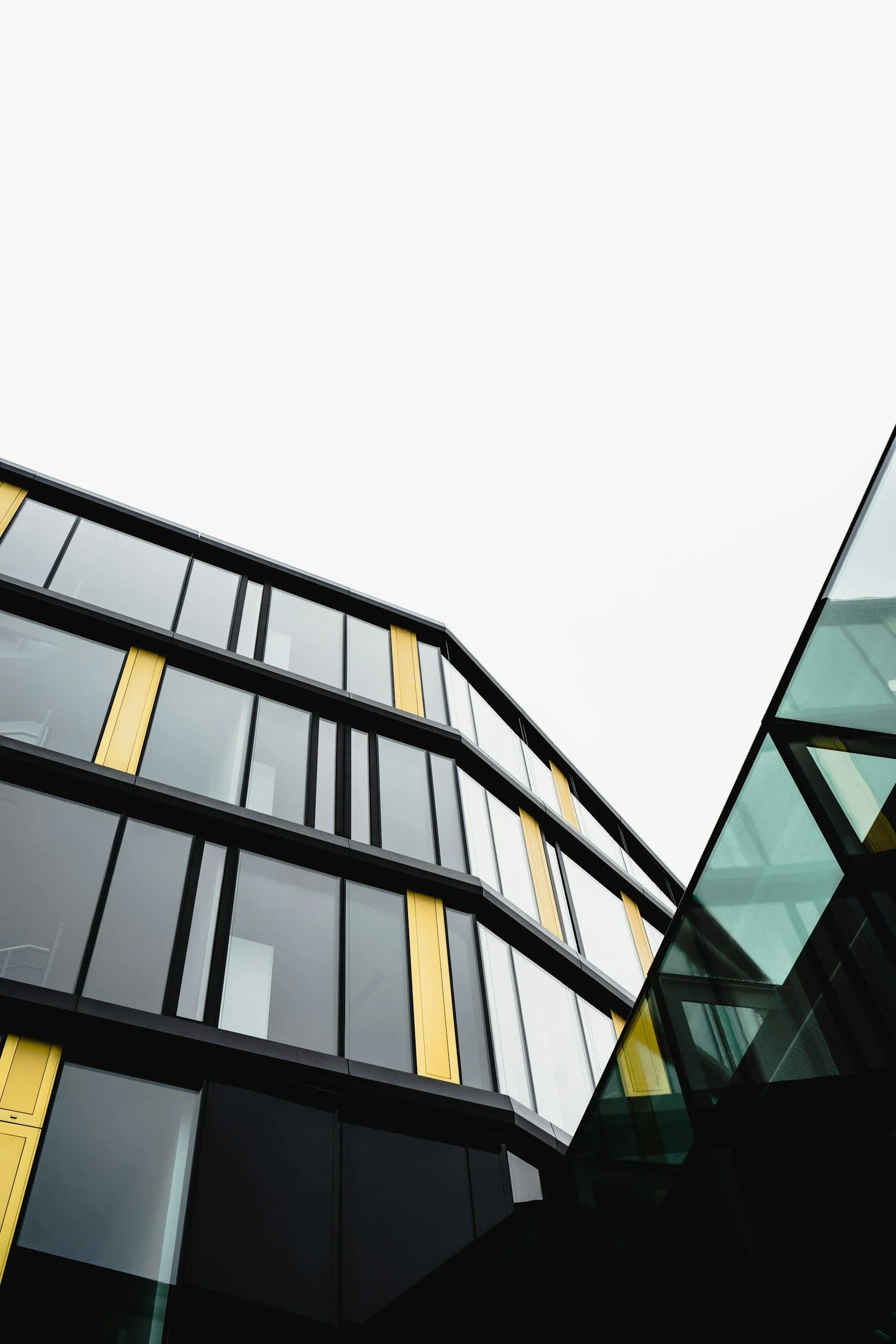 Low-angle view of a modern building with glass windows and yellow frames, set against an overcast sky.