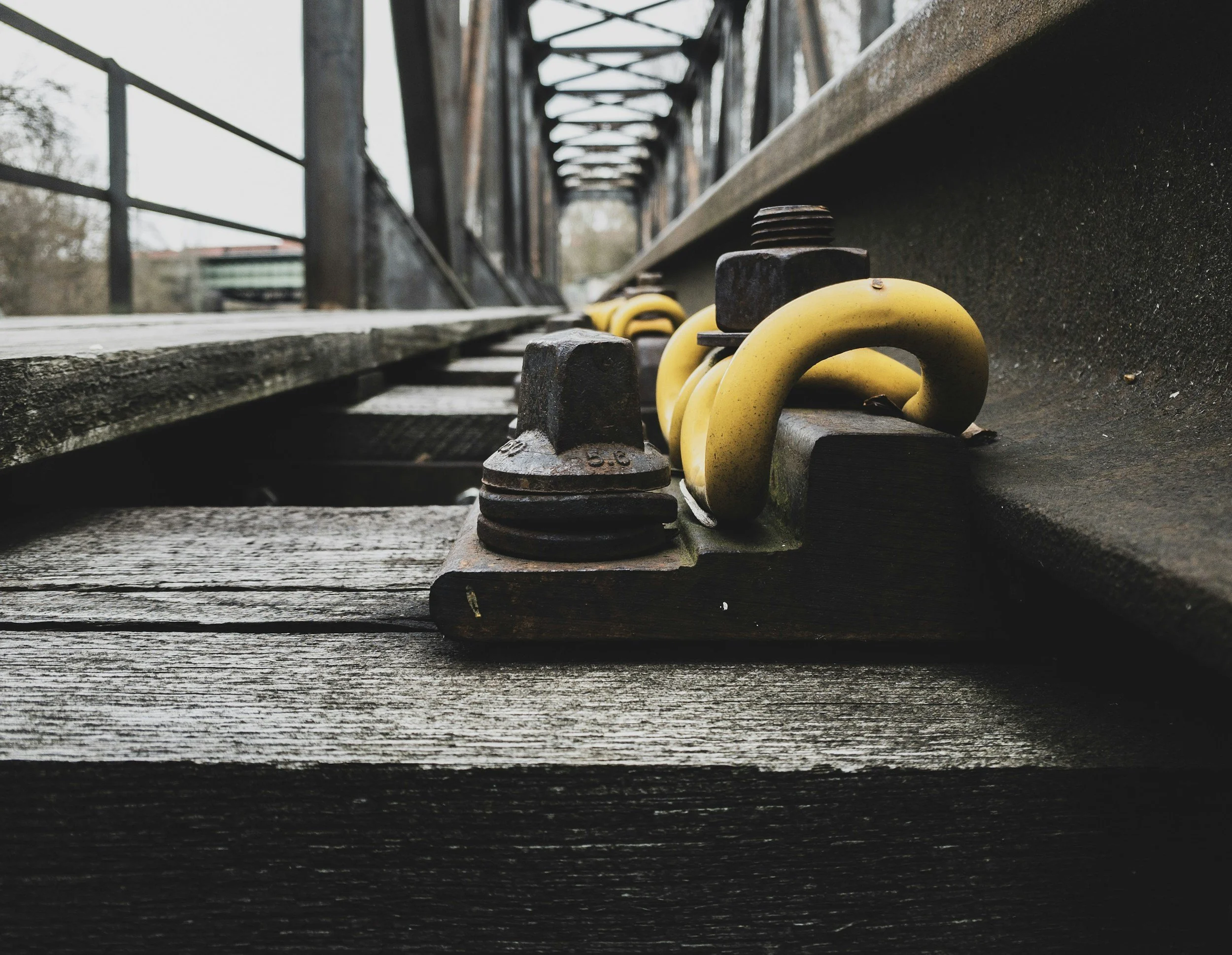 Close-up view of railroad equipment including yellow safety hooks and rusty bolts, on a weathered wooden railroad track with a steel bridge structure in the background.