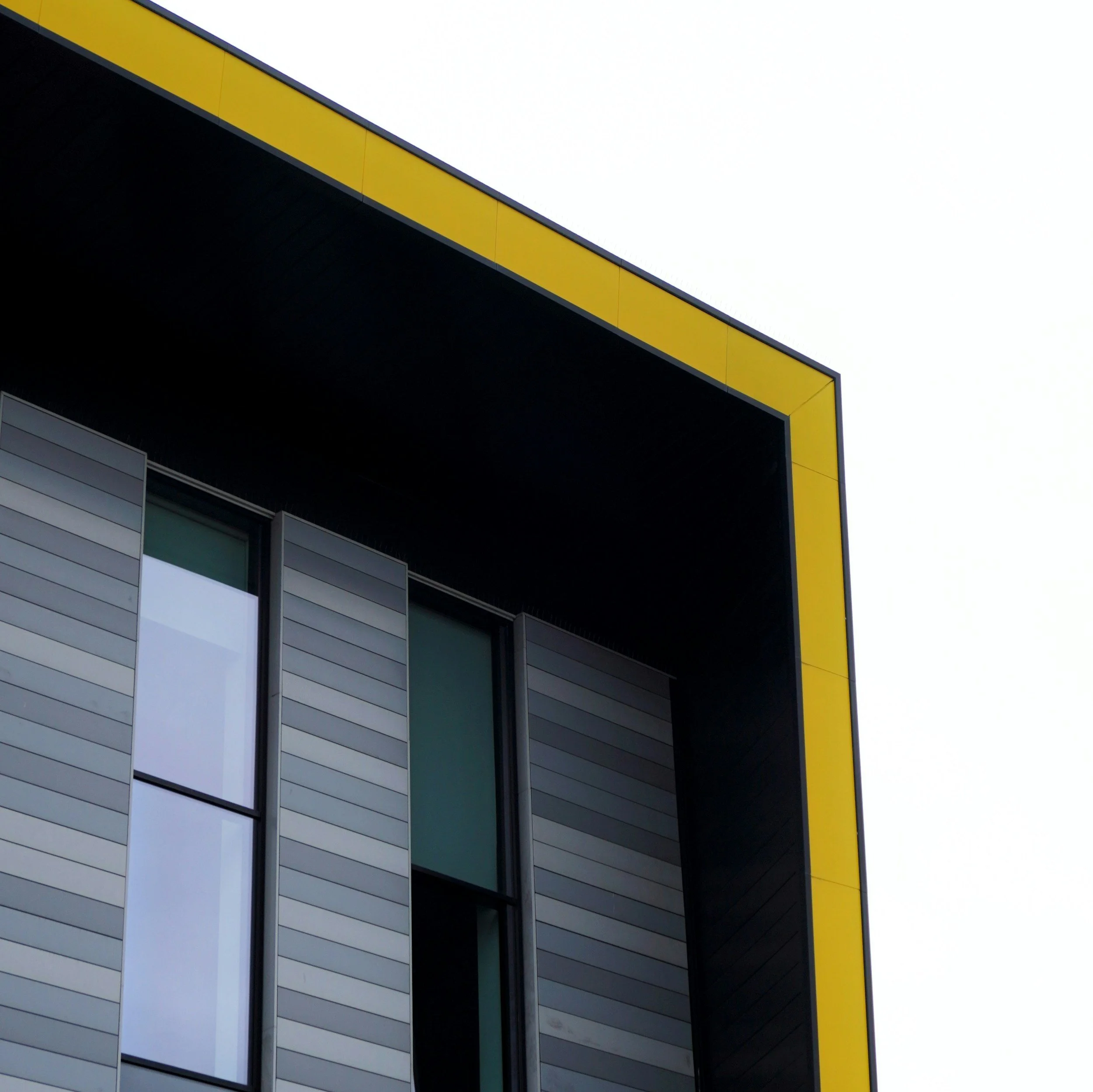 Close-up of a modern building's exterior with horizontal gray siding, large window panes, and a yellow accent border against a cloudy or overcast sky.