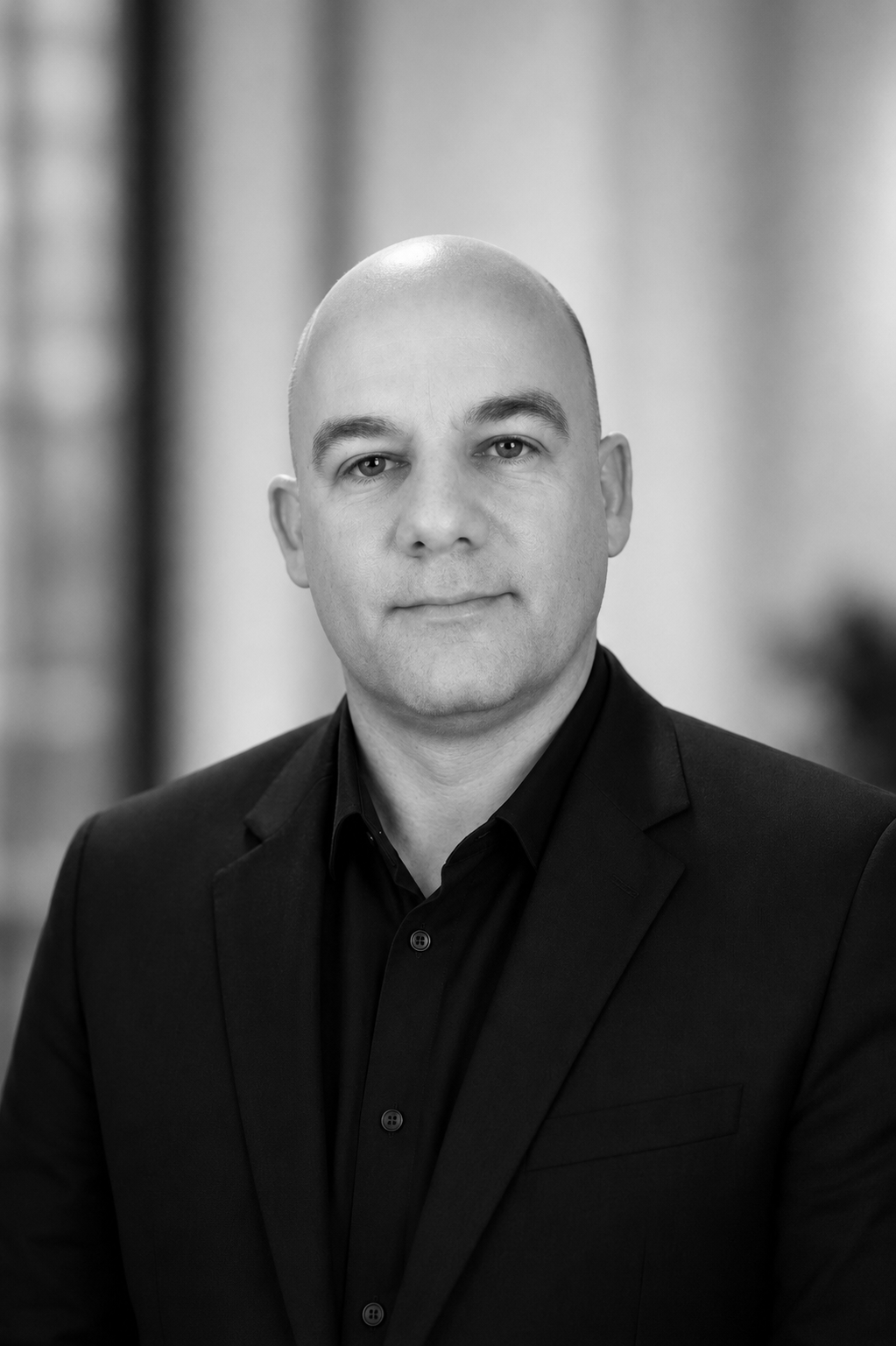 Black and white portrait of a bald man in a suit and black shirt, looking confidently at the camera with blurred background.