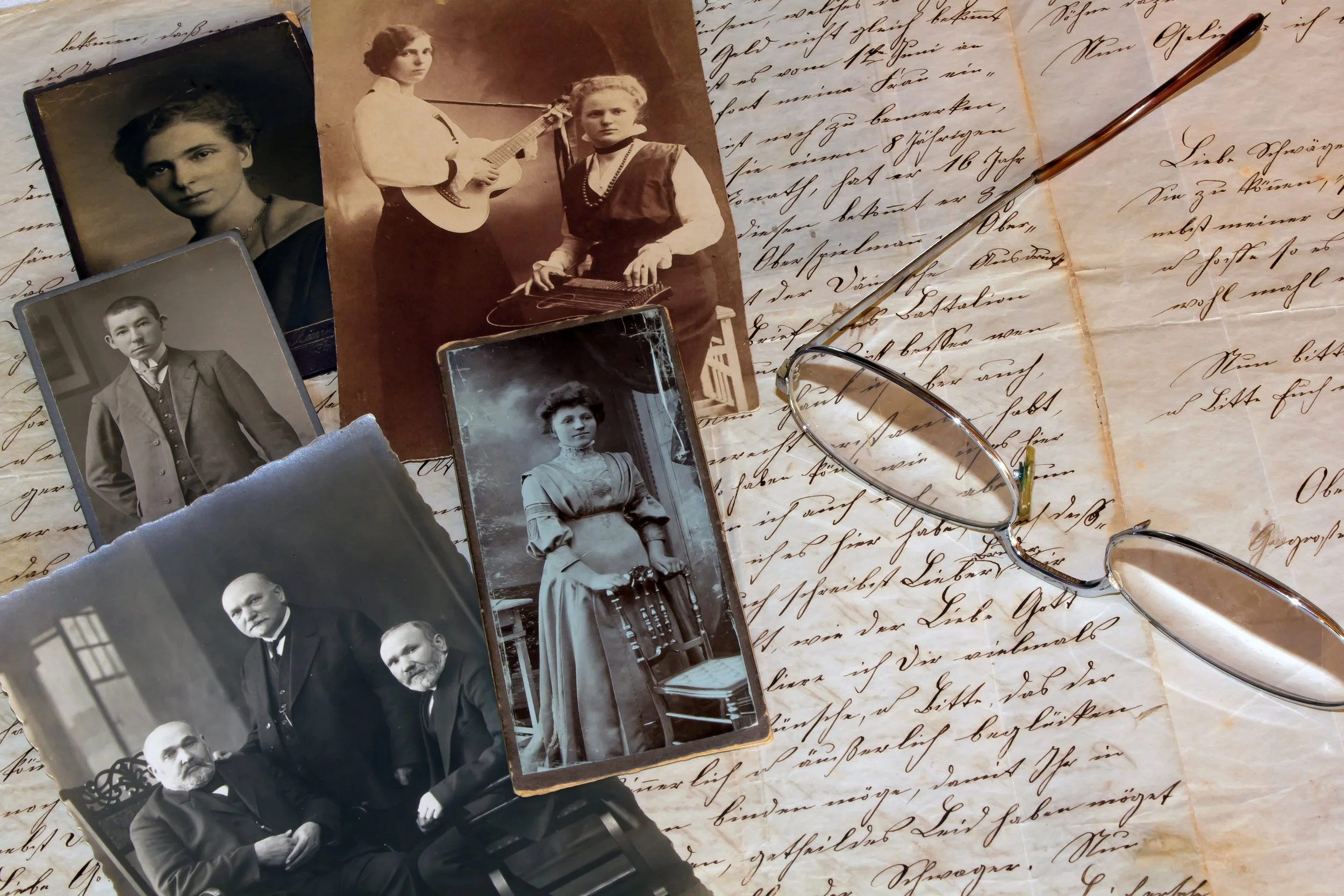 A collection of vintage black-and-white and sepia photographs of people, including a woman playing a guitar, a young boy, and a man with a beard. The photos are placed on handwritten letter paper, with glasses and a pen lying on the paper.