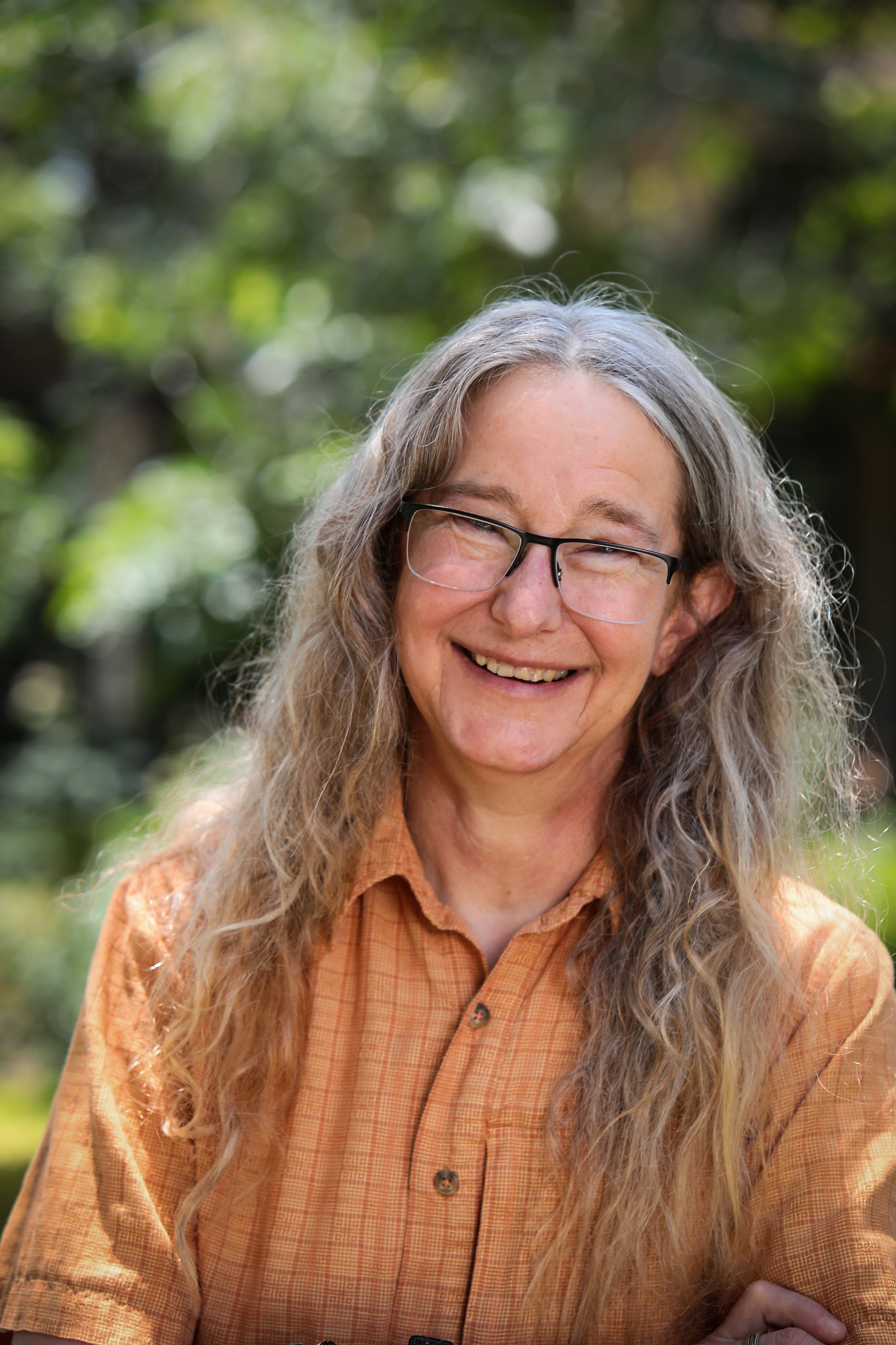 smiling woman with long, curly gray hair and glasses, wearing an orange checked shirt outdoors with green blurred background