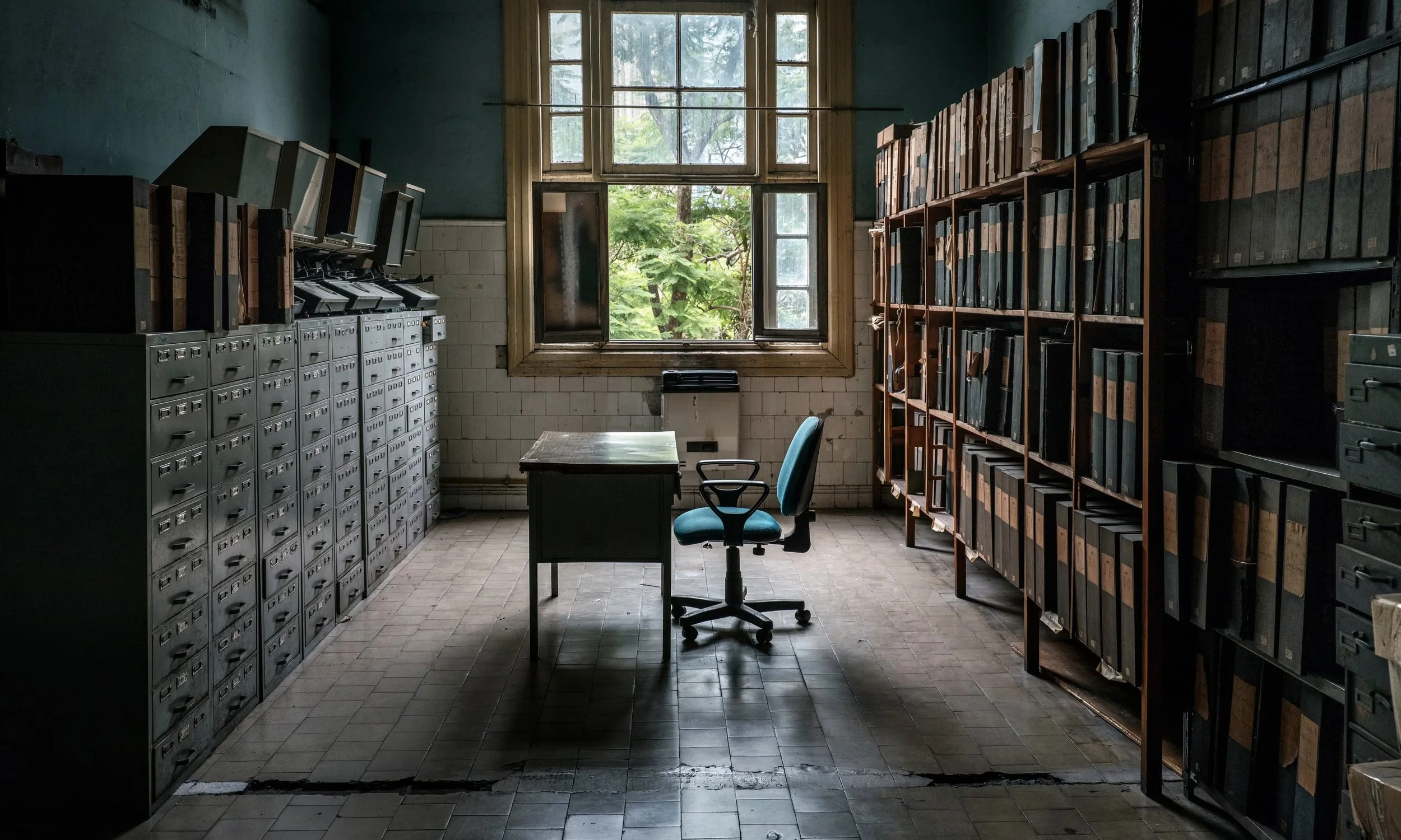 An old office with metal filing cabinets on the left, wooden bookshelves filled with binders on the right, a wooden desk in the center with a teal office chair, large window in the background revealing green trees outside, and aged tiled floor.