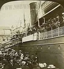Historical black and white photo of a large steamship docked at a pier with people on the dock and aboard, loading or unloading cargo.