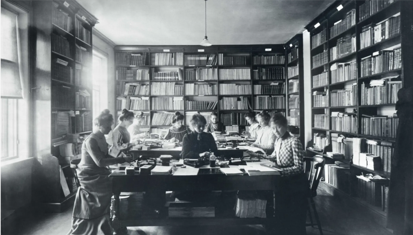 Black and white photo of a group of women working in a library surrounded by tall bookshelves filled with books, sitting around a large table.