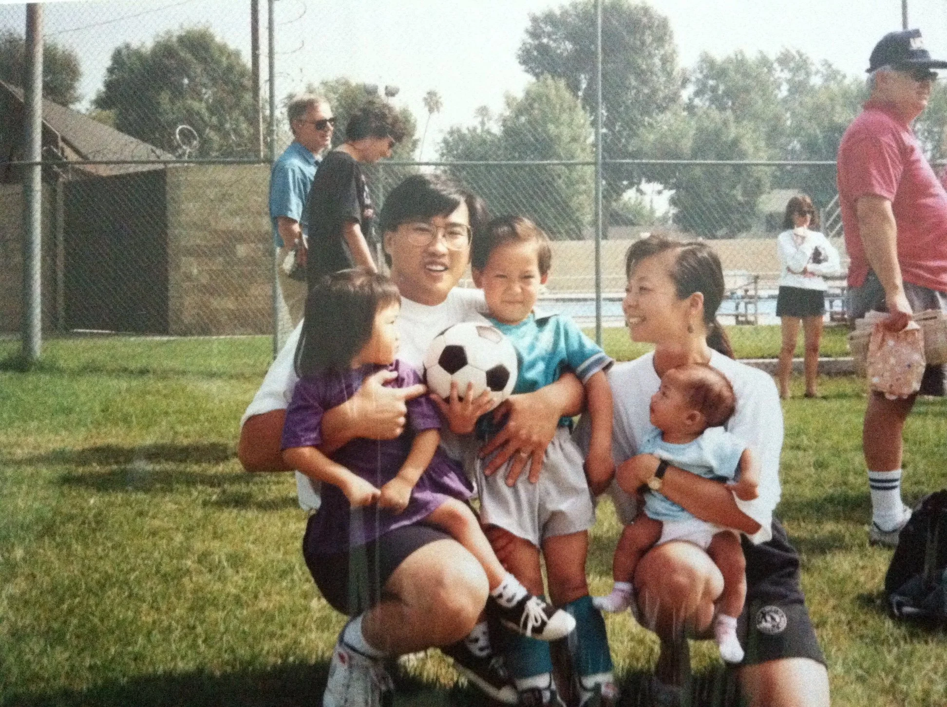 A group of children and adults on a grassy area at a park or sports field. Three children are held in the arms of a woman sitting on the grass, holding a soccer ball. The children are smiling and appear happy. Several adults are standing in the backg