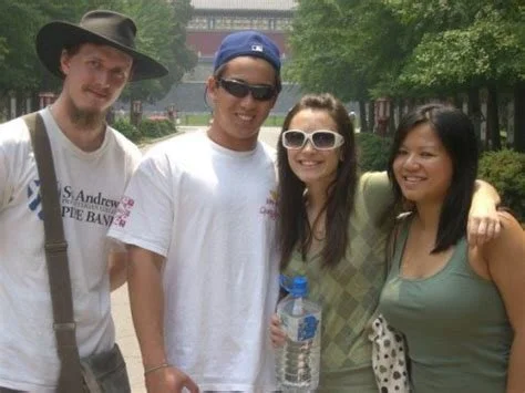Group of four young friends standing outdoors, smiling, with a park and trees in the background, dressed casually.