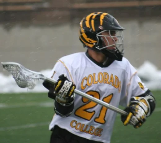 Lacrosse player in white jersey with yellow and black helmet holding a lacrosse stick on the field.