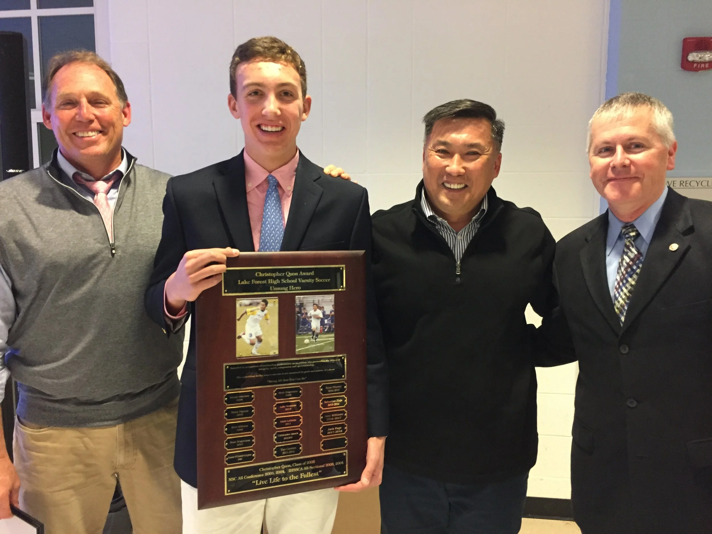Four men standing together indoors, with the second man from the left holding a framed award plaque with a photograph of a soccer player and text. The man on the far right is wearing a suit and tie, the others are in casual or semi-formal attire. The background includes a white wall, a black object, and a small red sign.